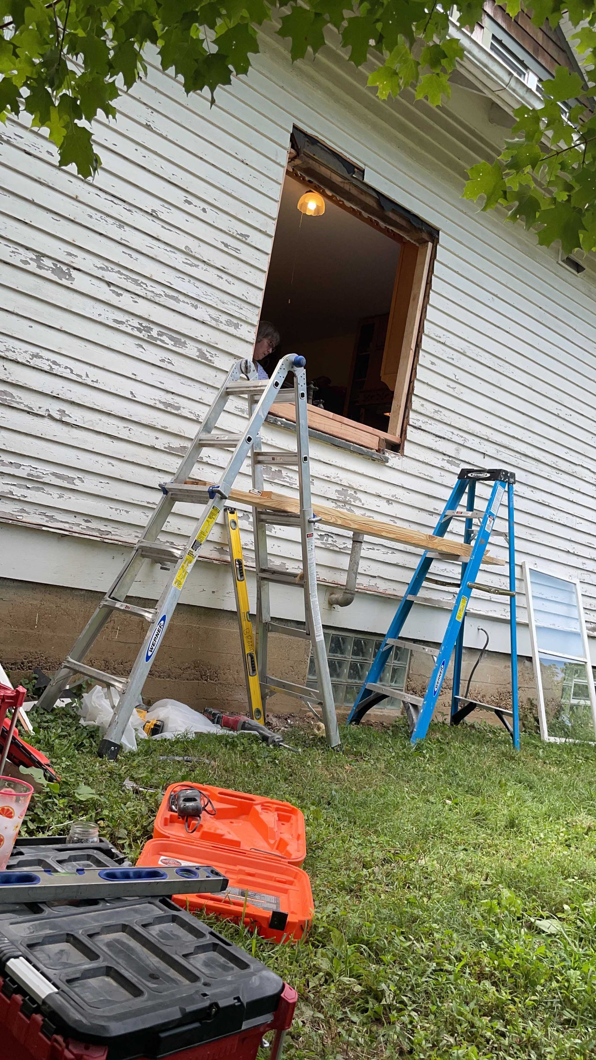 Ladder propped against a house with an open window. Construction tools and equipment are on the ground.