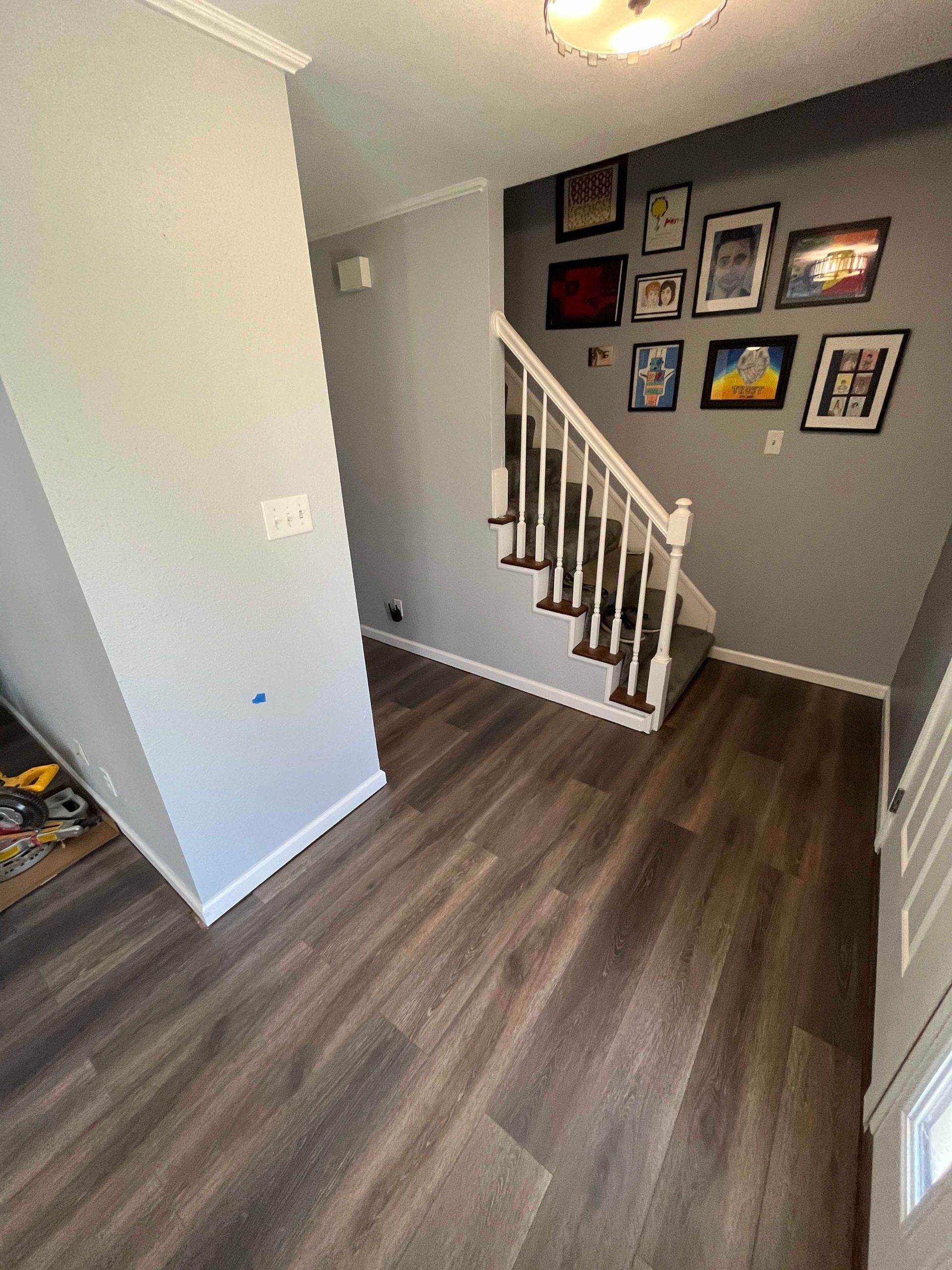 Entryway with wood-look floor, stairs, and framed photos on a gray wall.