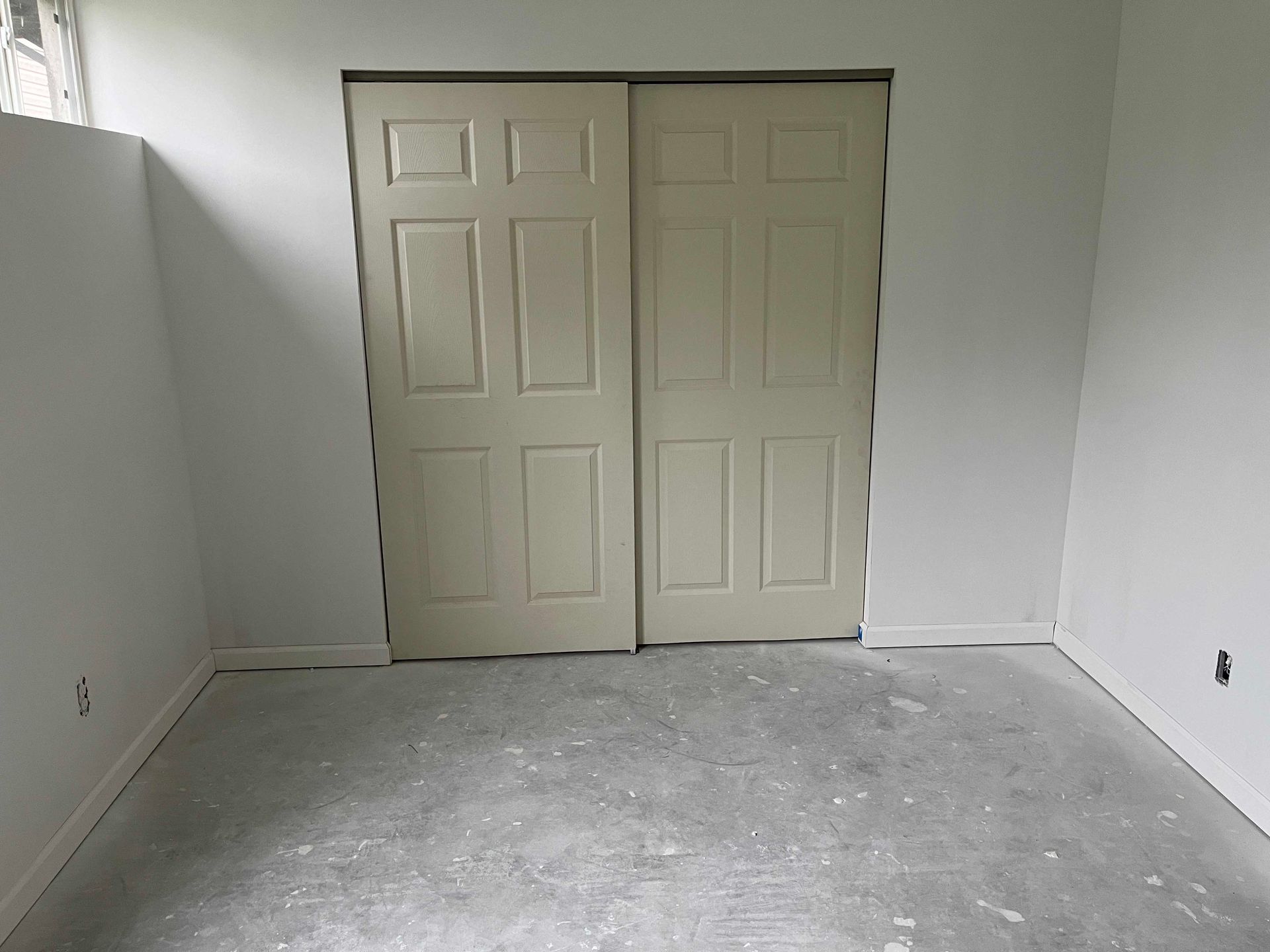 Interior view of a room with two beige sliding closet doors. Walls are white, floor is concrete.