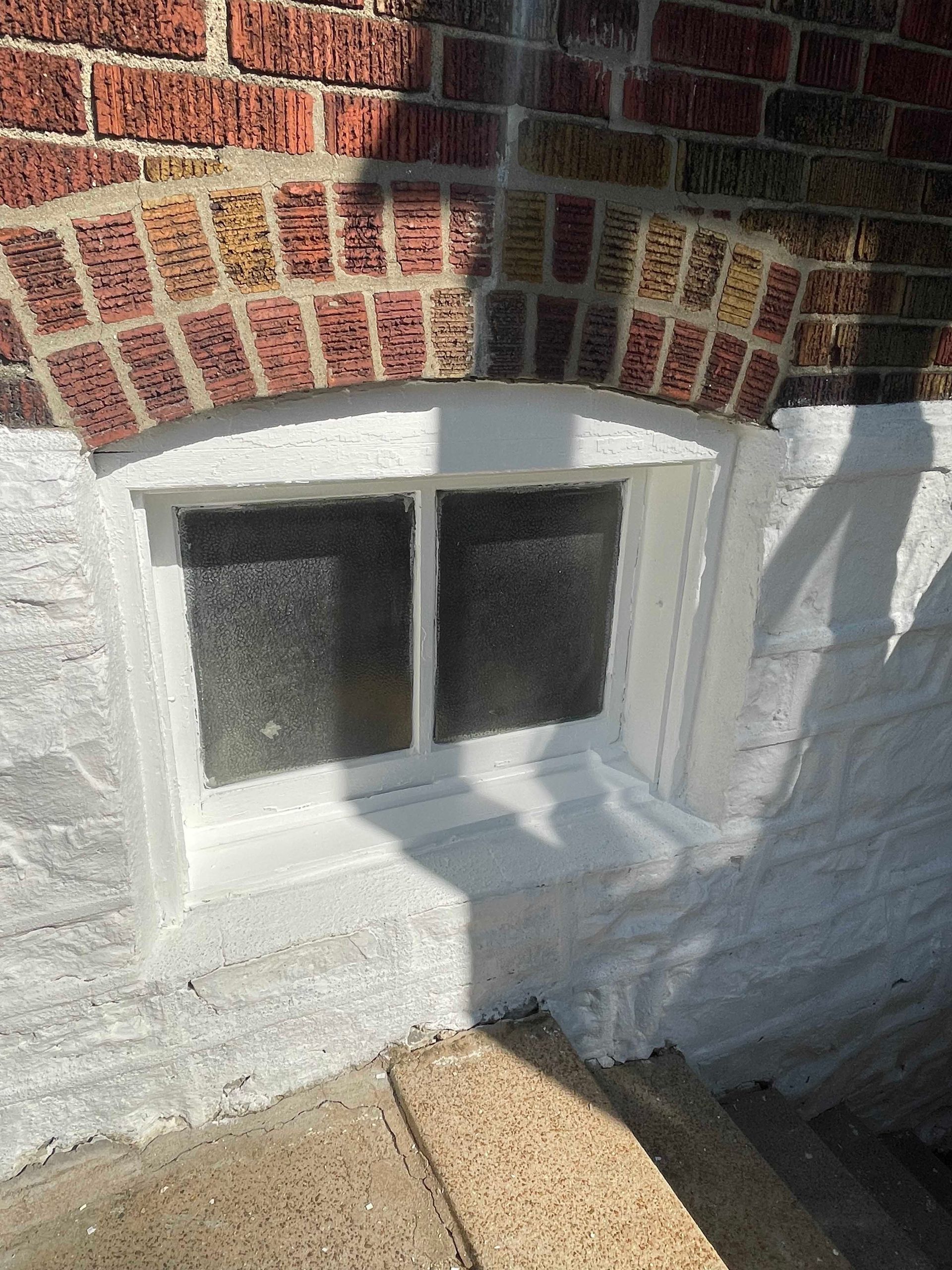 White-framed basement window in brick wall. Concrete steps lead to the window. Bright sunlight casts a shadow.