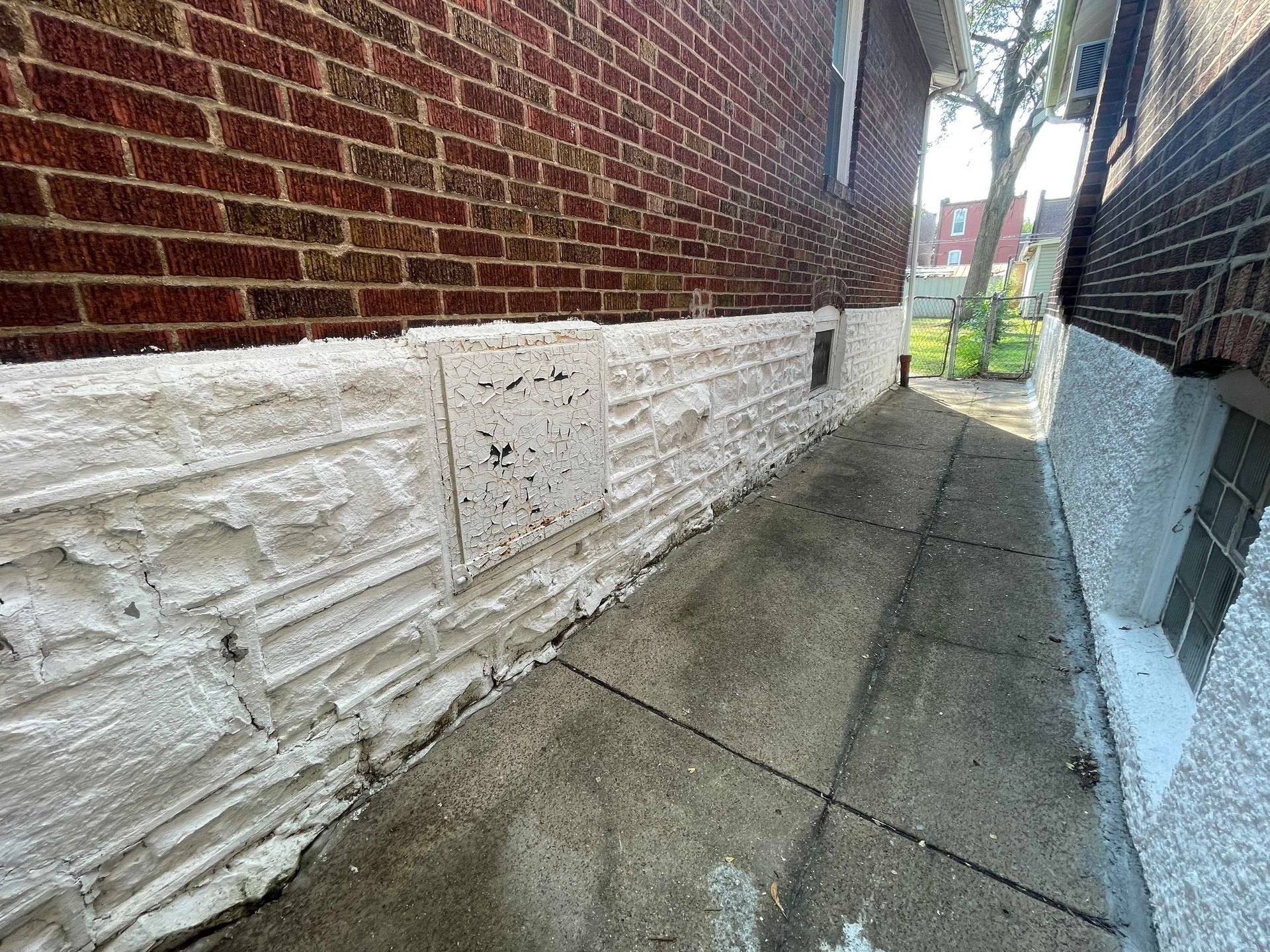 Exterior view of a narrow alleyway with a brick building and a textured, white-painted concrete wall alongside a sidewalk.