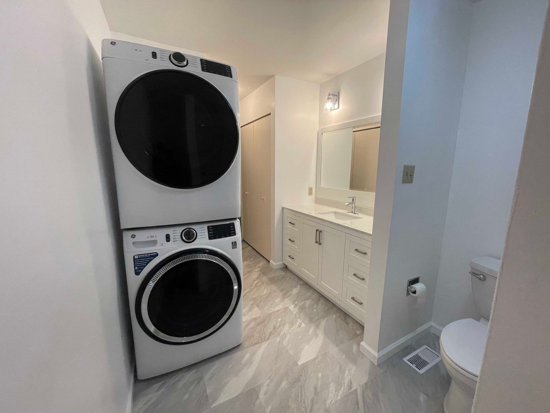 Stacked white washer and dryer in a bathroom with white vanity, mirror, and toilet. Gray flooring.