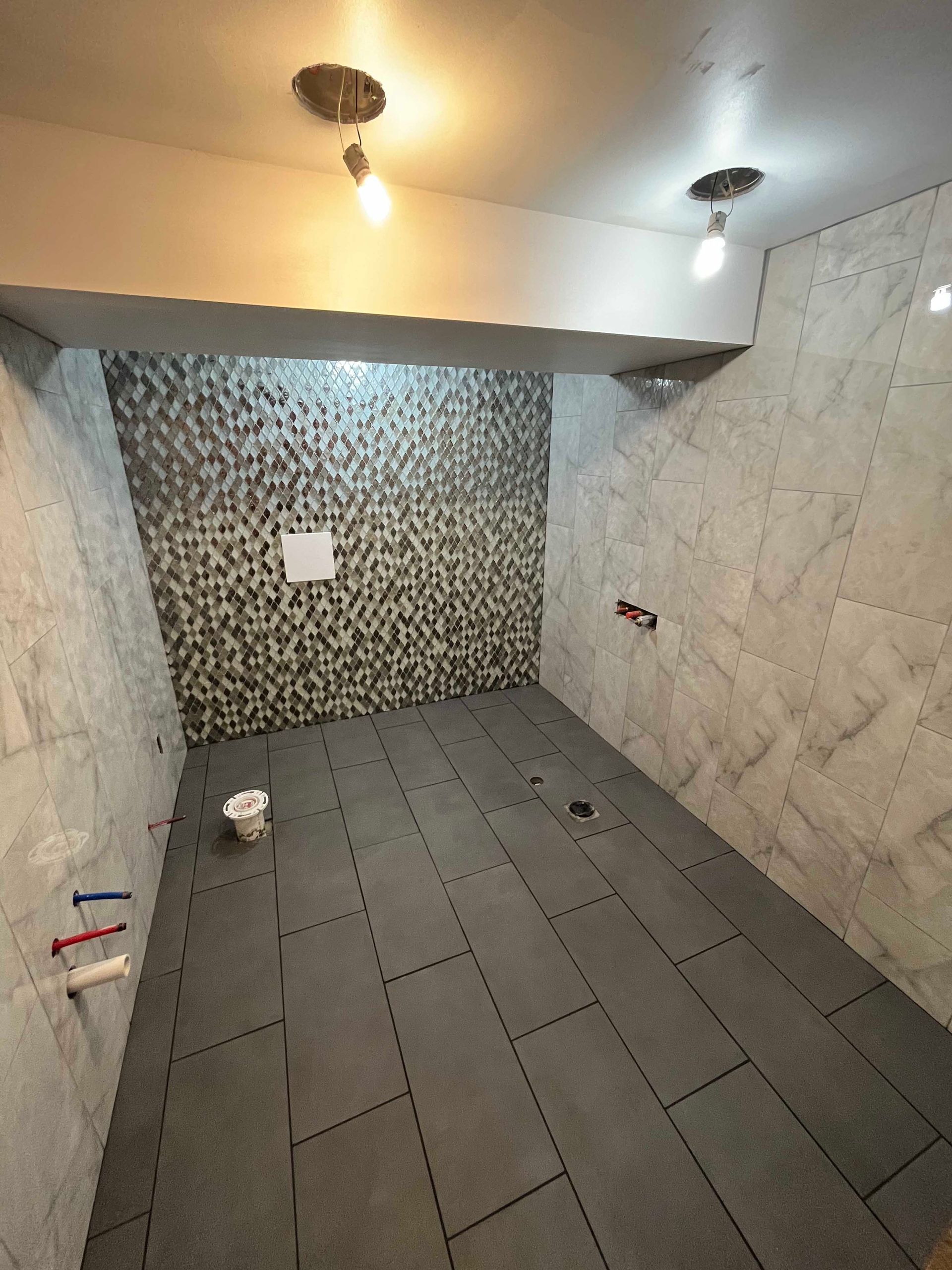 Bathroom renovation in progress. Gray tile floor, light gray wall tiles, textured mosaic accent wall, exposed plumbing.