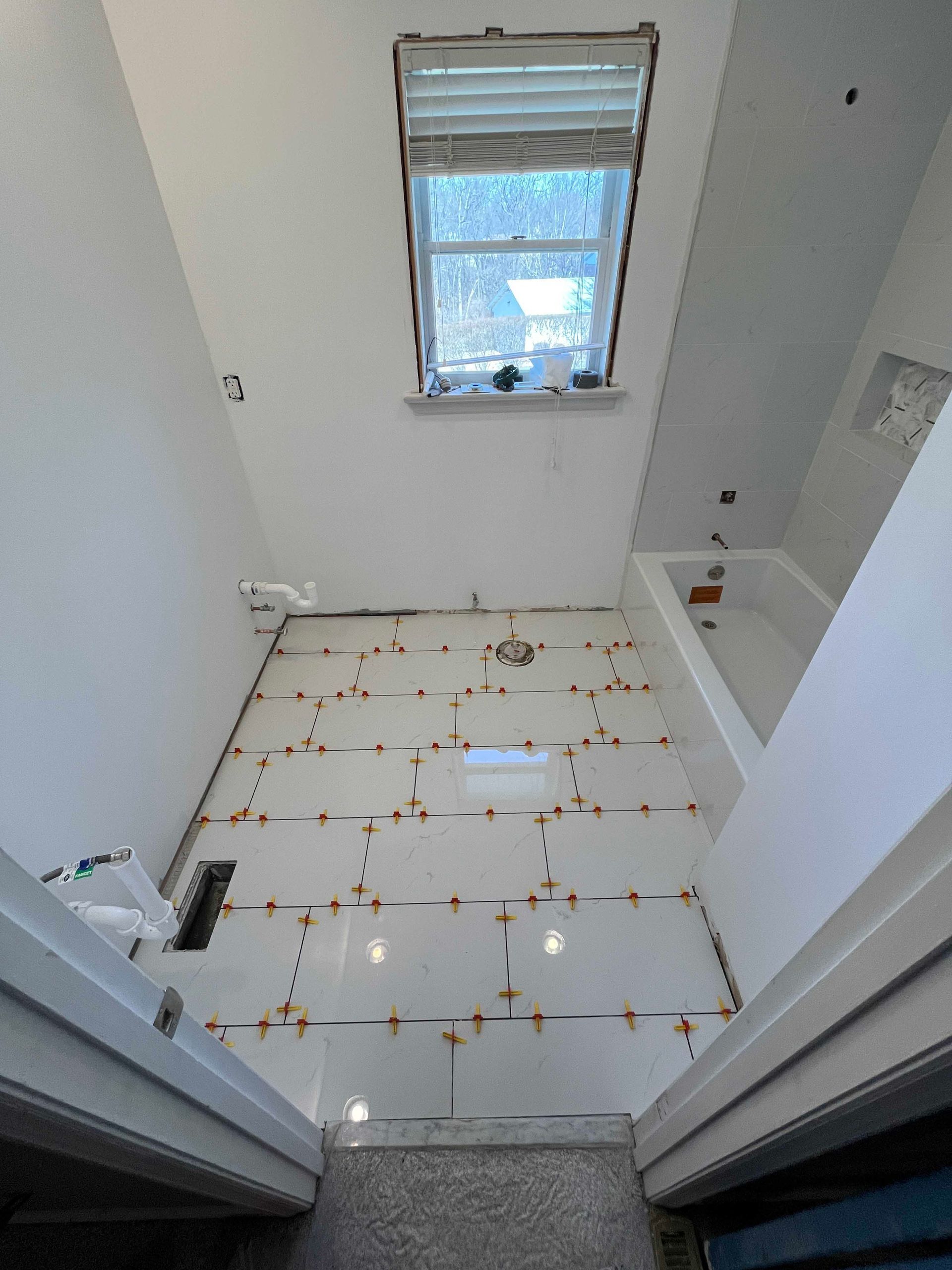 Bathroom under construction, white tiles on floor, window with blinds, white walls, and a bathtub.