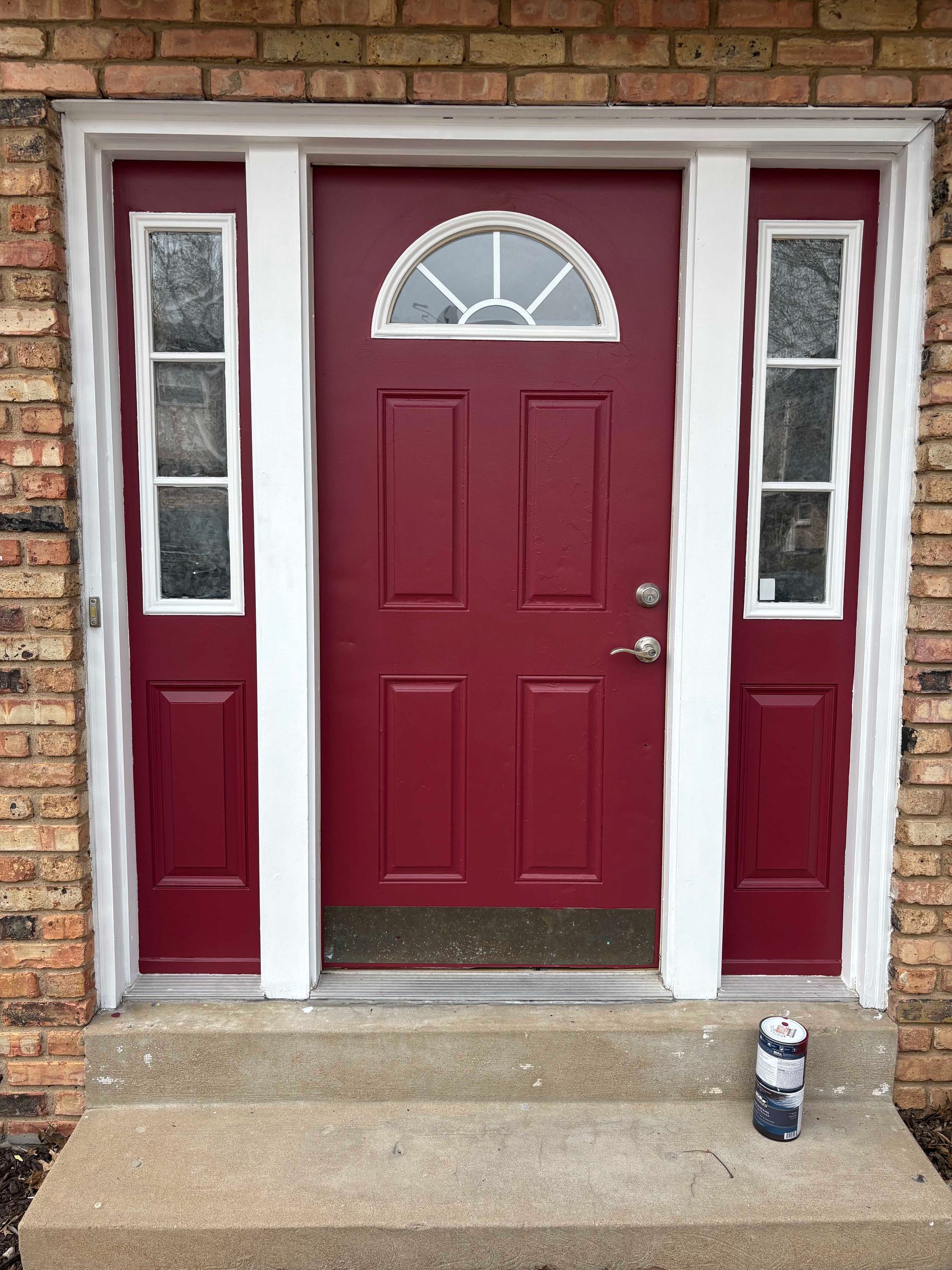 Red front door with sidelights, set in brick building with concrete steps.