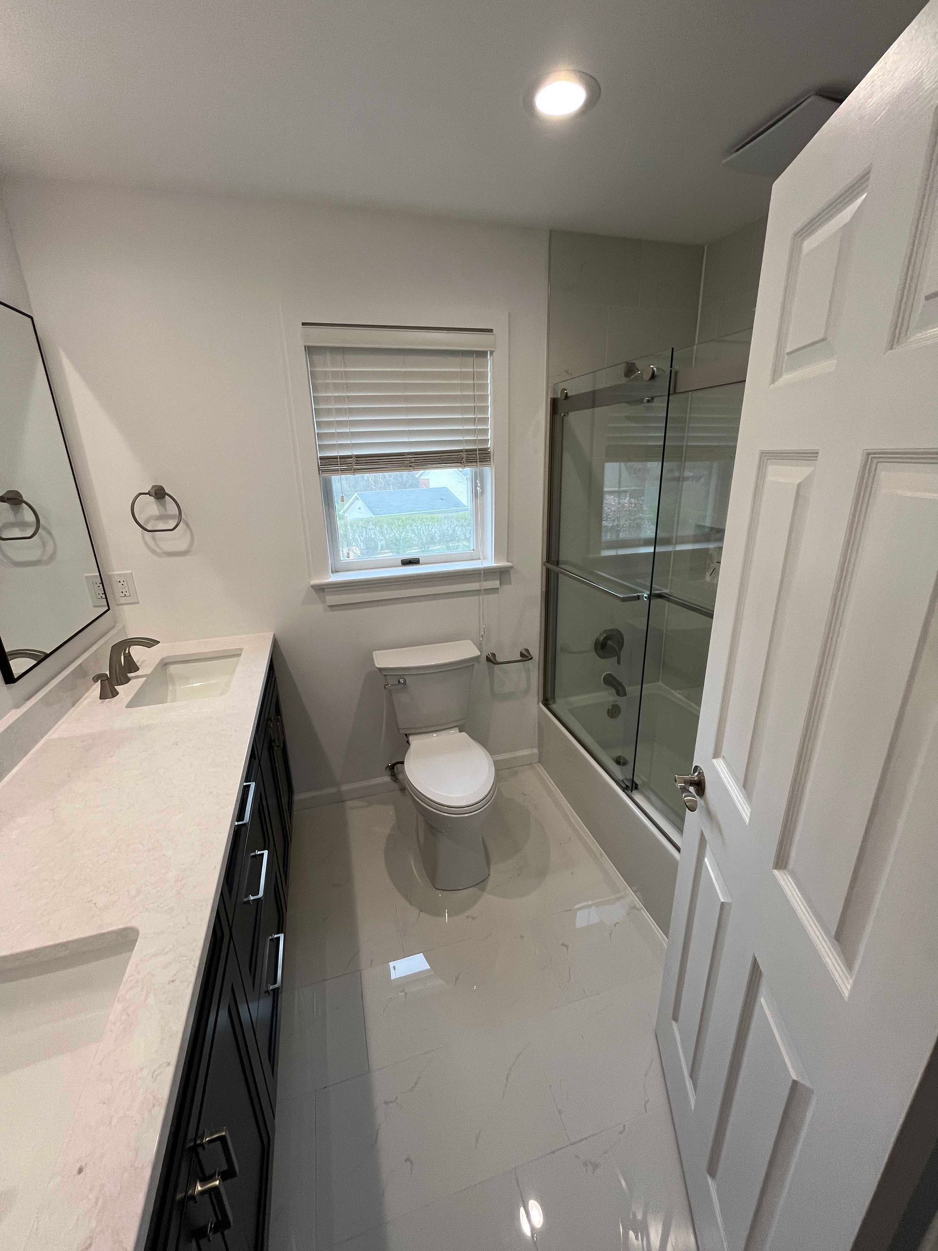 Bathroom with white walls, vanity, toilet, shower, and window.