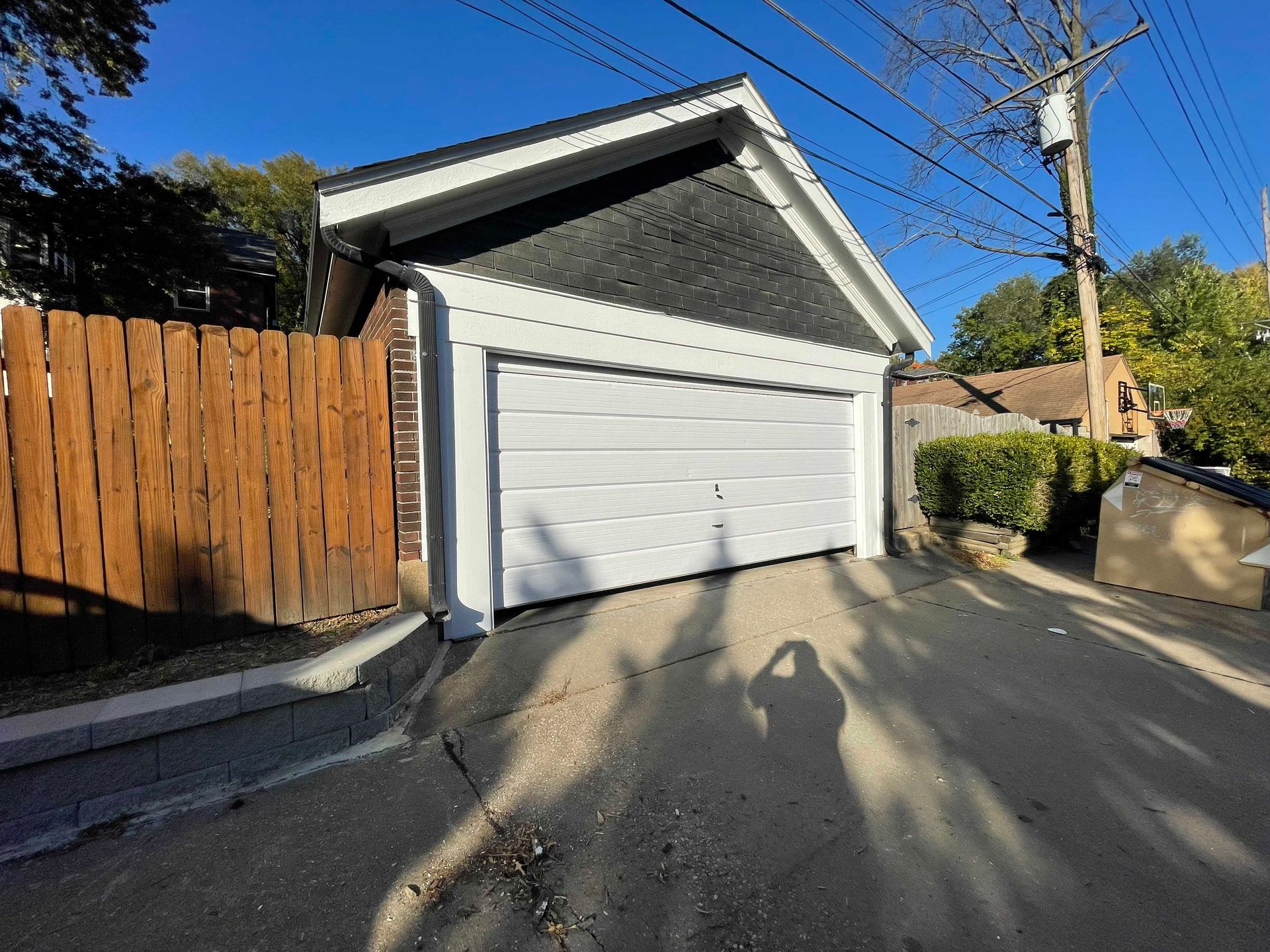 White garage with black roof, bordered by white trim and a wooden fence, on a sunny day.