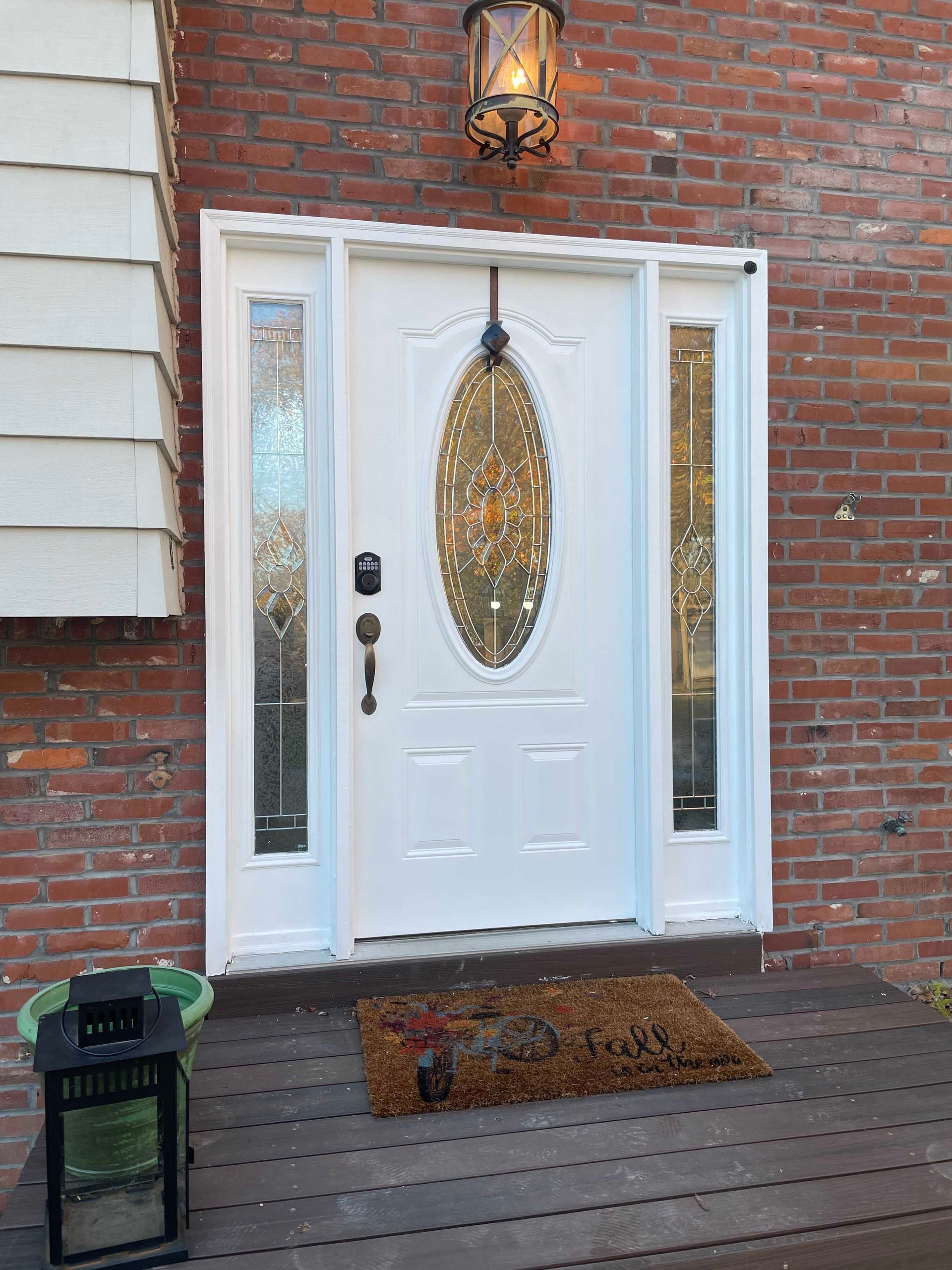 White front door with oval glass panel and sidelights, set in brick building. A doormat and lantern are at the base.