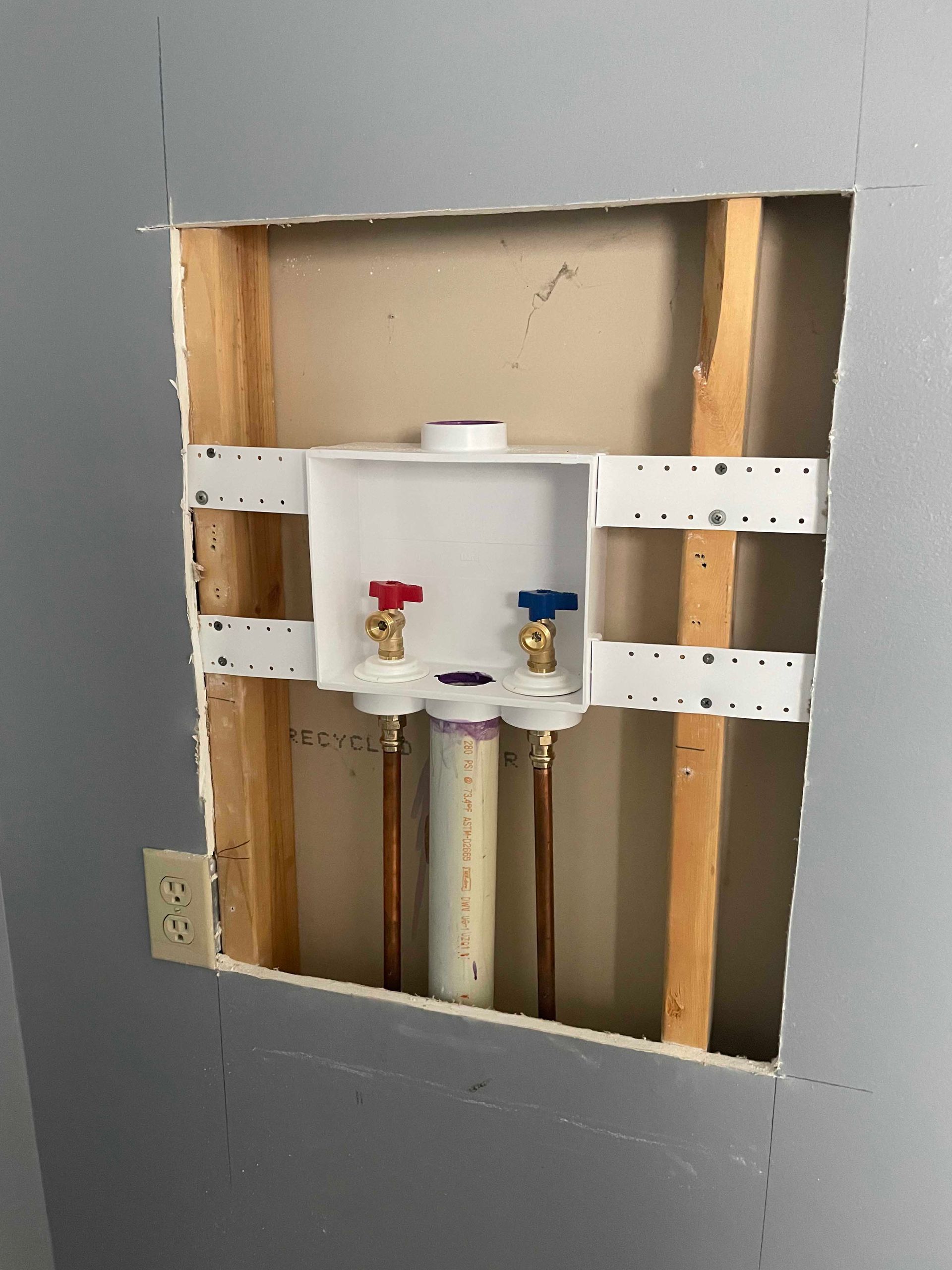 Laundry room plumbing: a white box with water valves, copper pipes, and a drainpipe installed within a wall cutout.