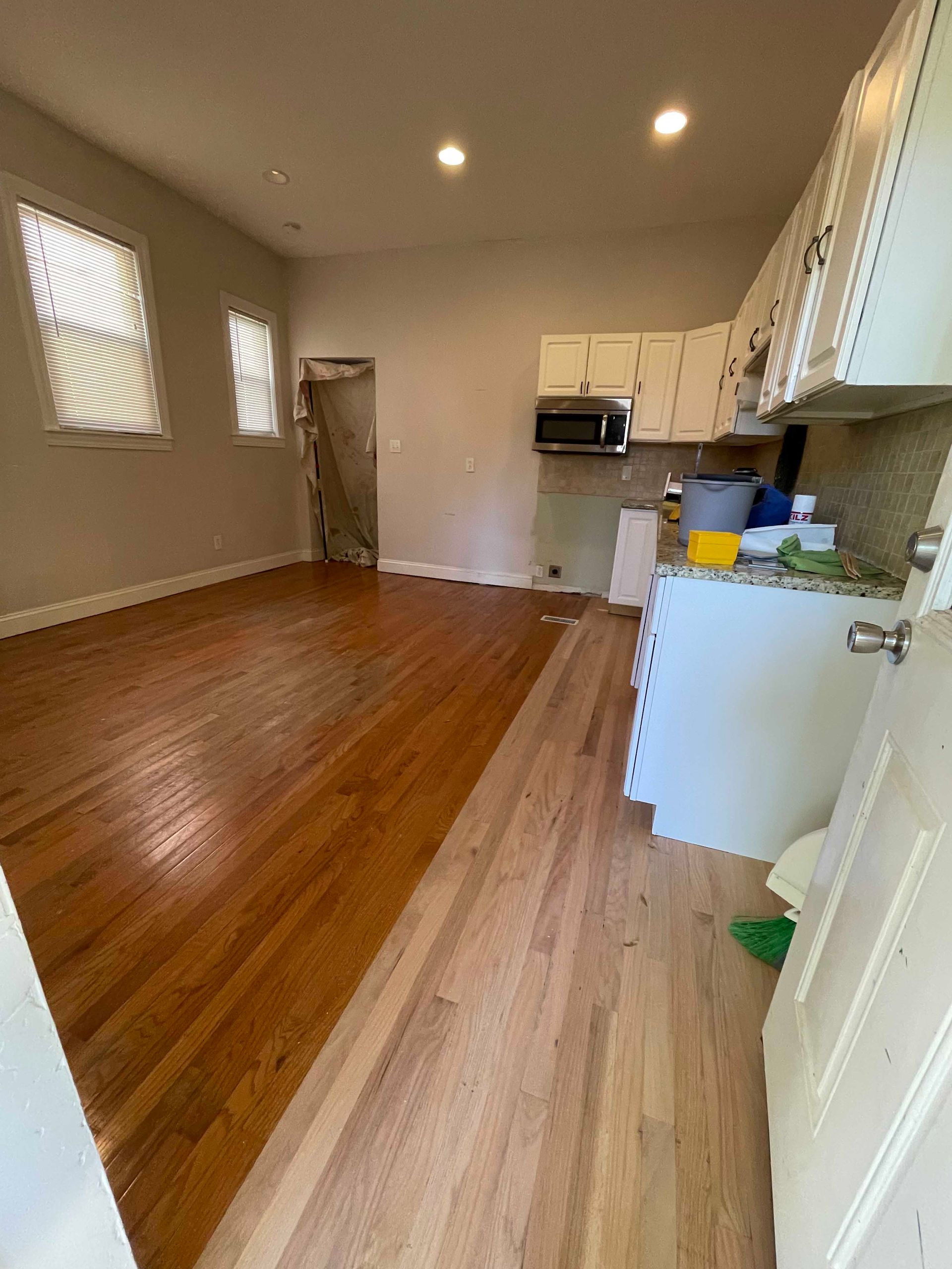 Open-concept living space with hardwood floors, kitchen cabinets, and two windows.