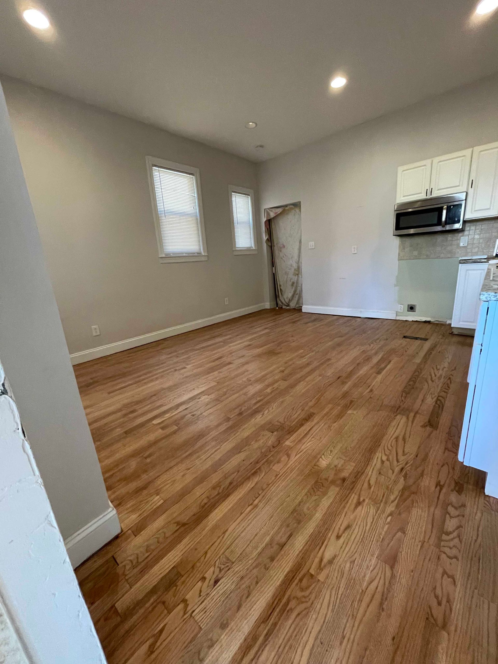 Empty room with wood floors, neutral walls, two windows, and kitchen cabinets.