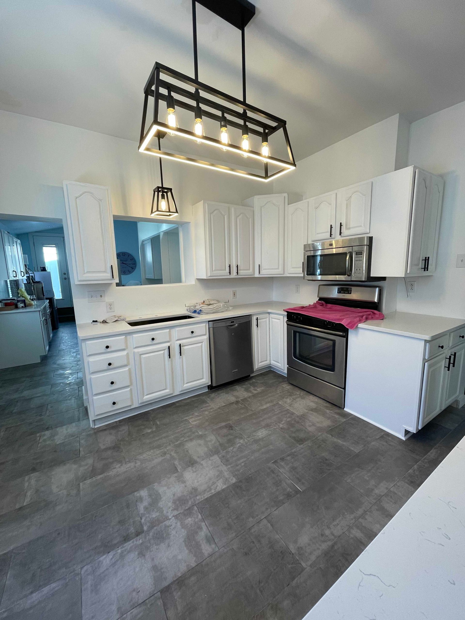 Modern kitchen with white cabinets, stainless steel appliances, and gray tiled floor, a large window, and rectangular light fixture.