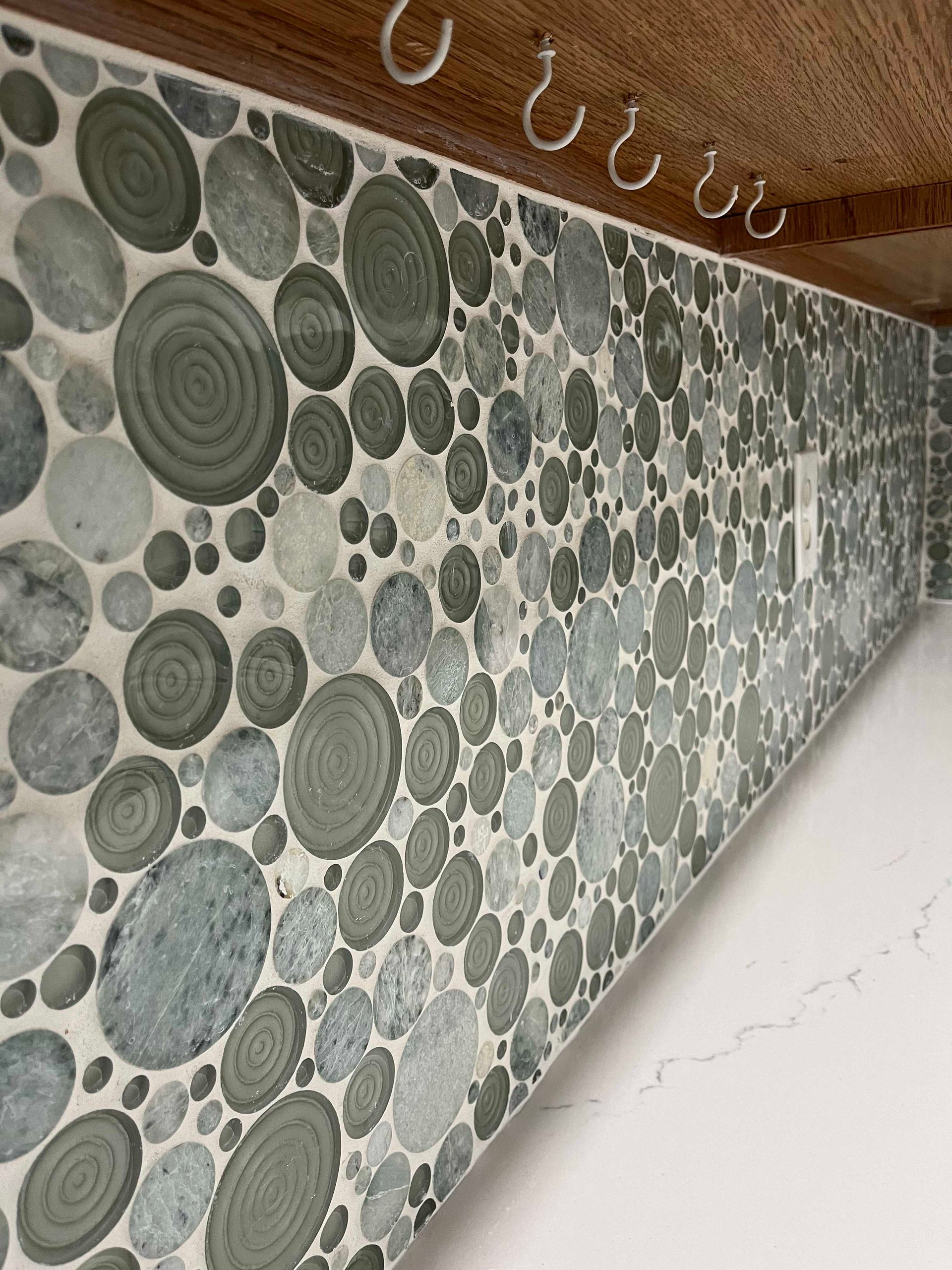 Tiled backsplash with circular green and gray stone pattern above a white countertop, with hooks.
