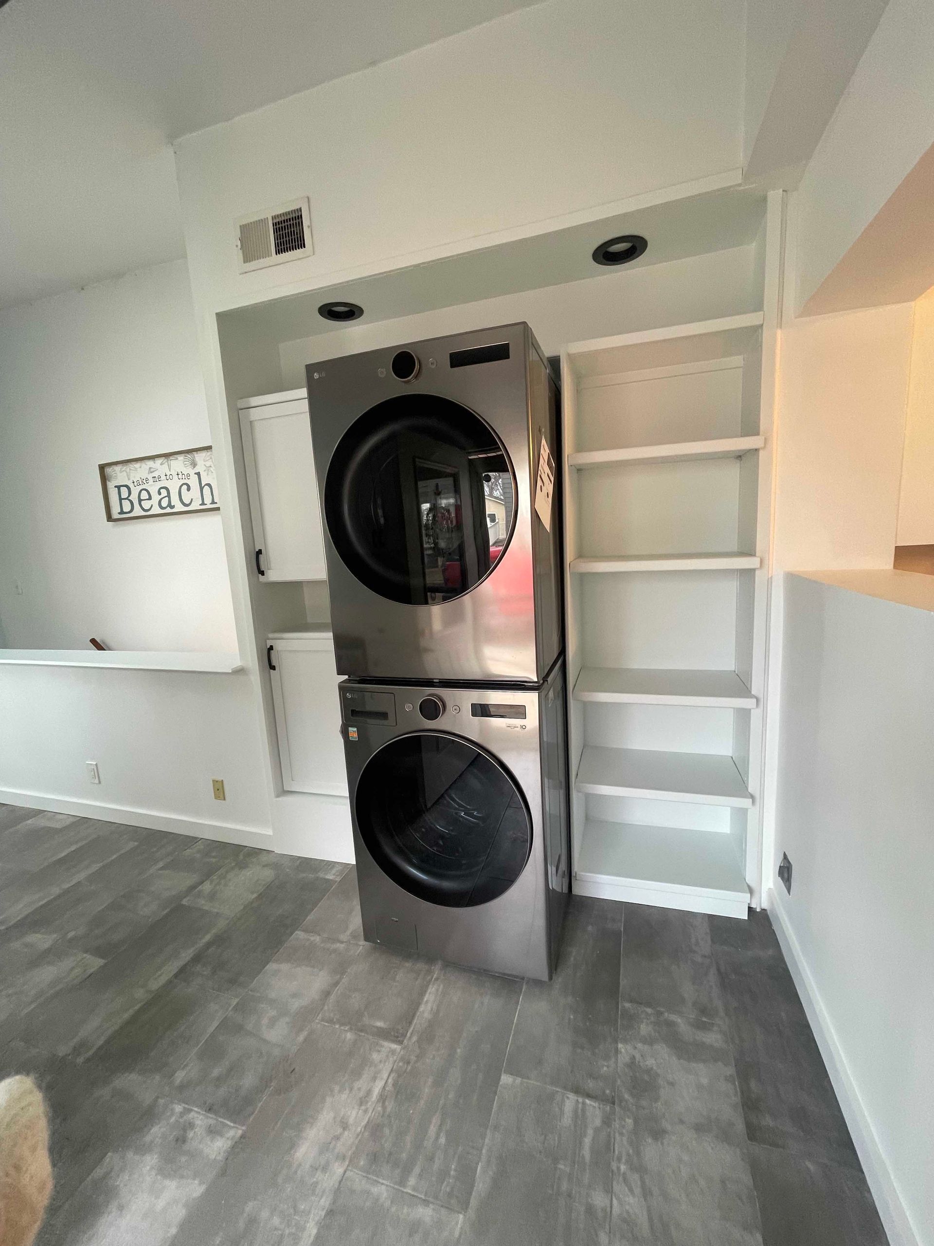 Stacked washer and dryer in a white alcove, next to white shelving. Gray flooring.
