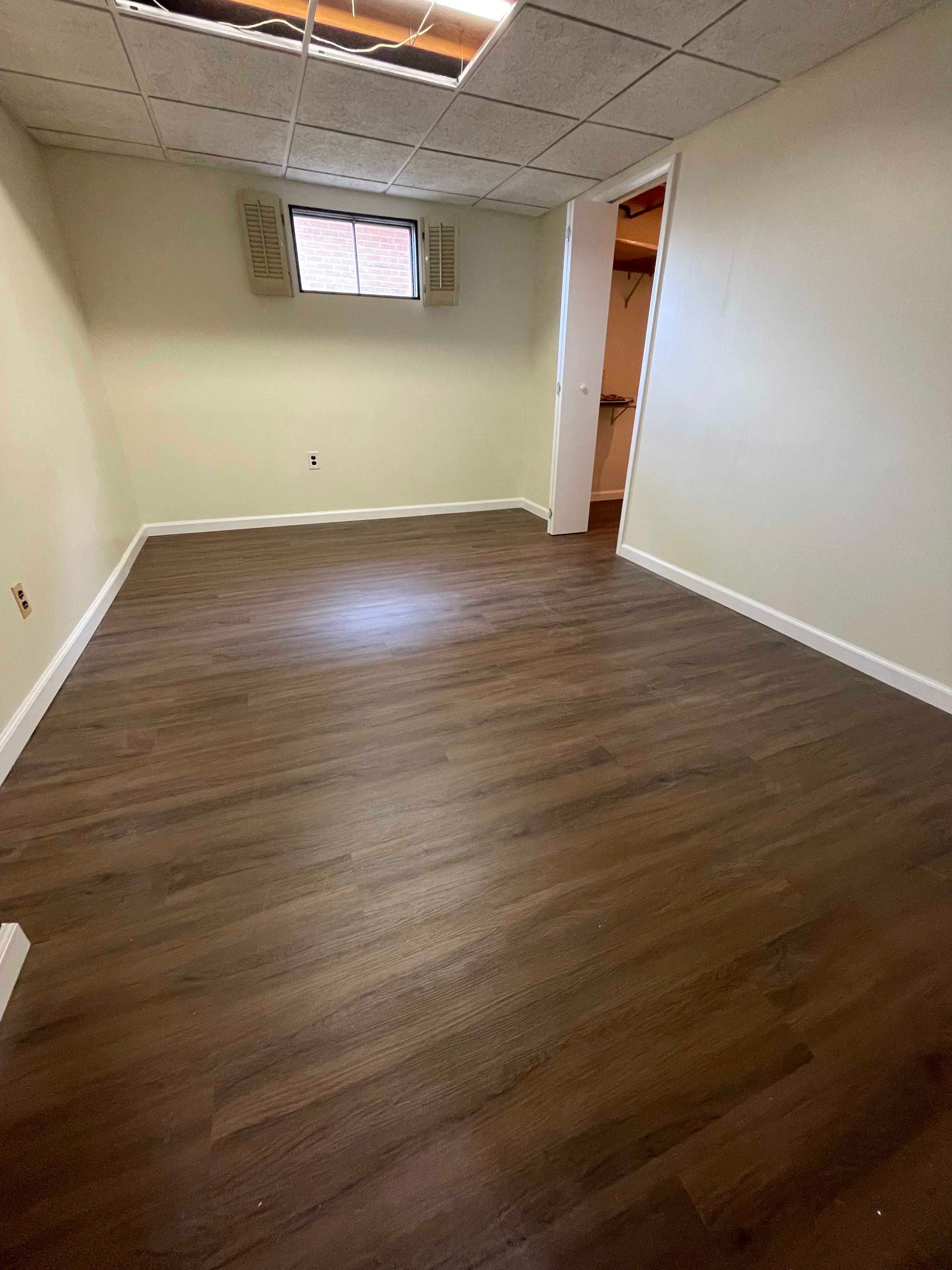 Empty room with brown flooring, off-white walls, a small window, and a doorway to a closet.