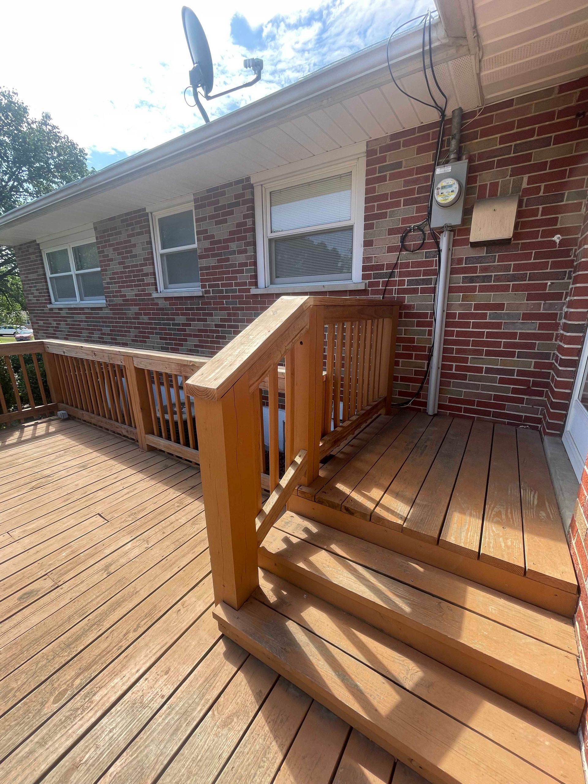 Wooden deck with stairs, railing, and brick house exterior. Satellite dish and electrical box visible.