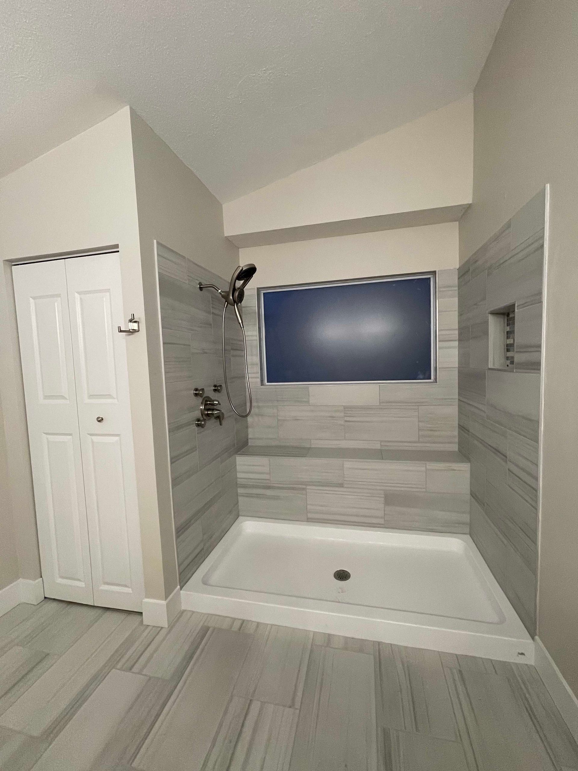 Bathroom with a tiled shower stall and a window. Light gray tile covers the walls and floor. A white door is visible.