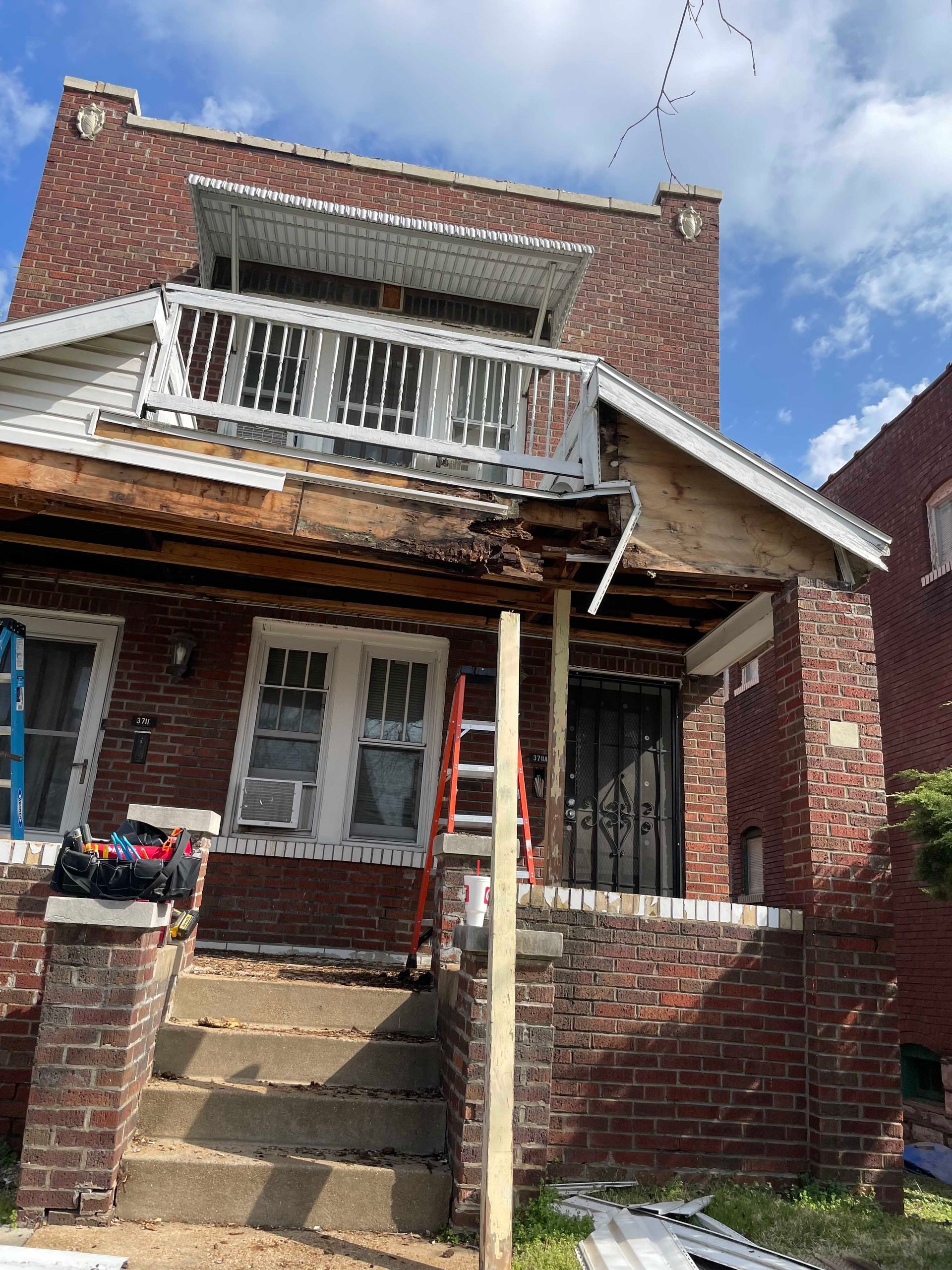 Brick house undergoing renovation, porch and second-floor balcony visible, with a ladder leaning against the porch.