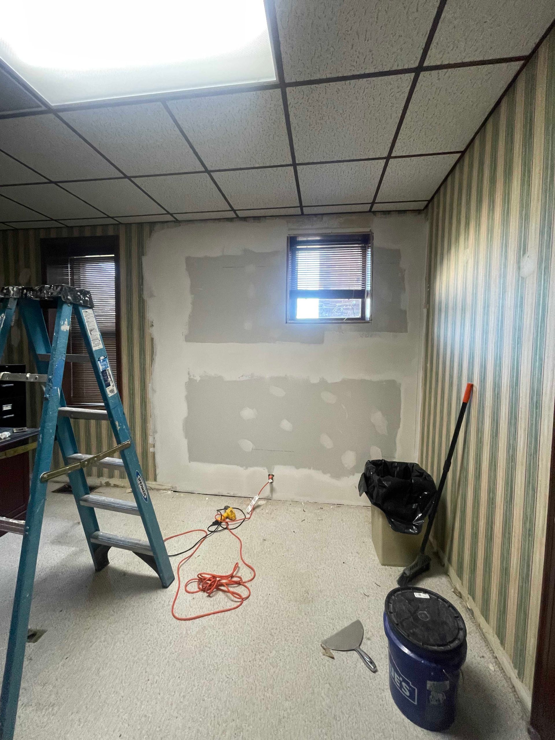 A room under renovation with exposed drywall patches and tools; ladder, trash bag, paint bucket.