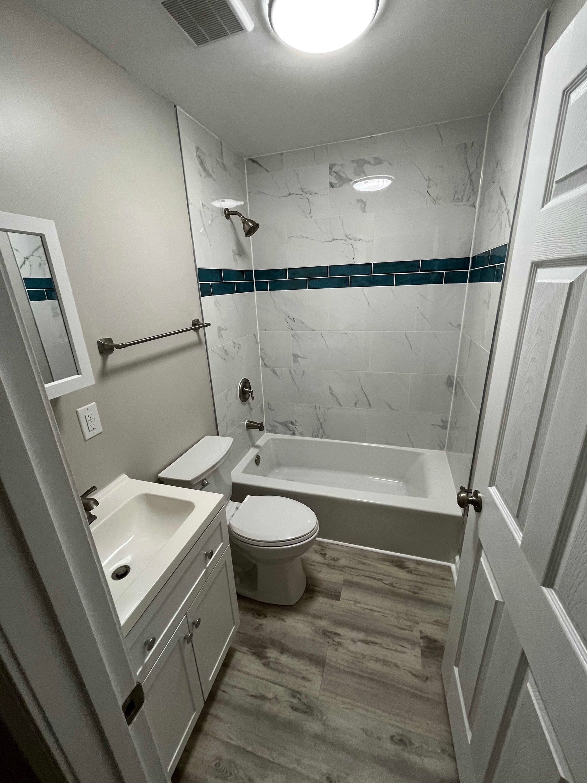 Bathroom with white fixtures, marble-look tiled shower, teal accent tile, and wood-look flooring.
