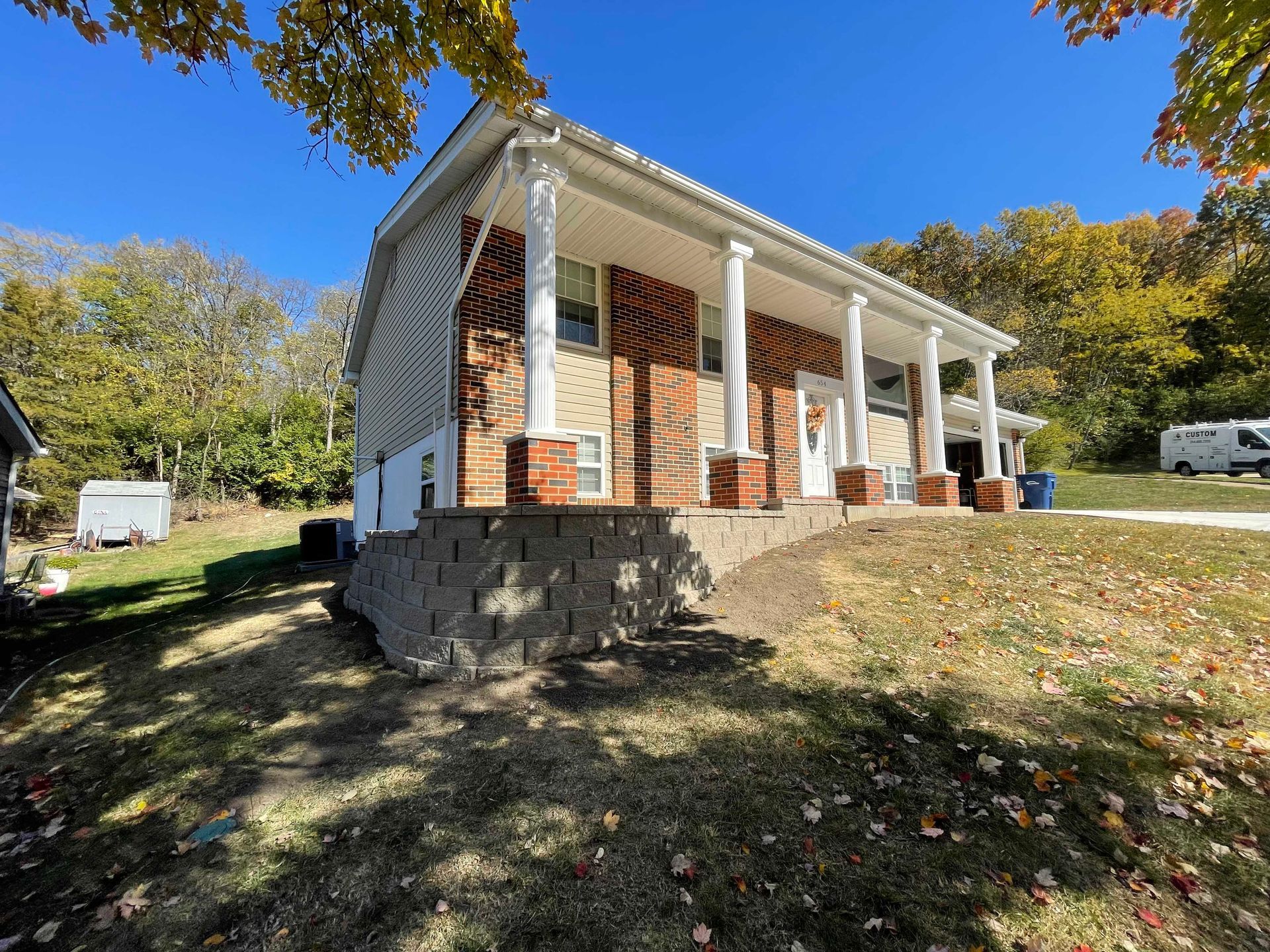 House with white columns, brick, and retaining wall on a grassy hillside under a blue sky.