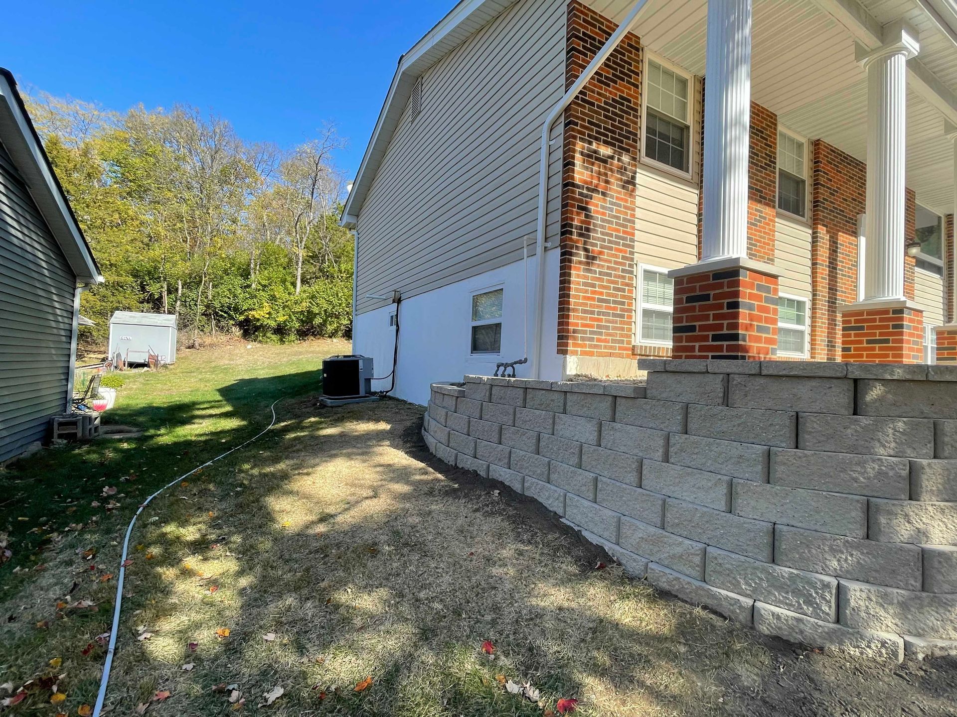 A two-story house with a brick and concrete block retaining wall and dry grass in front.