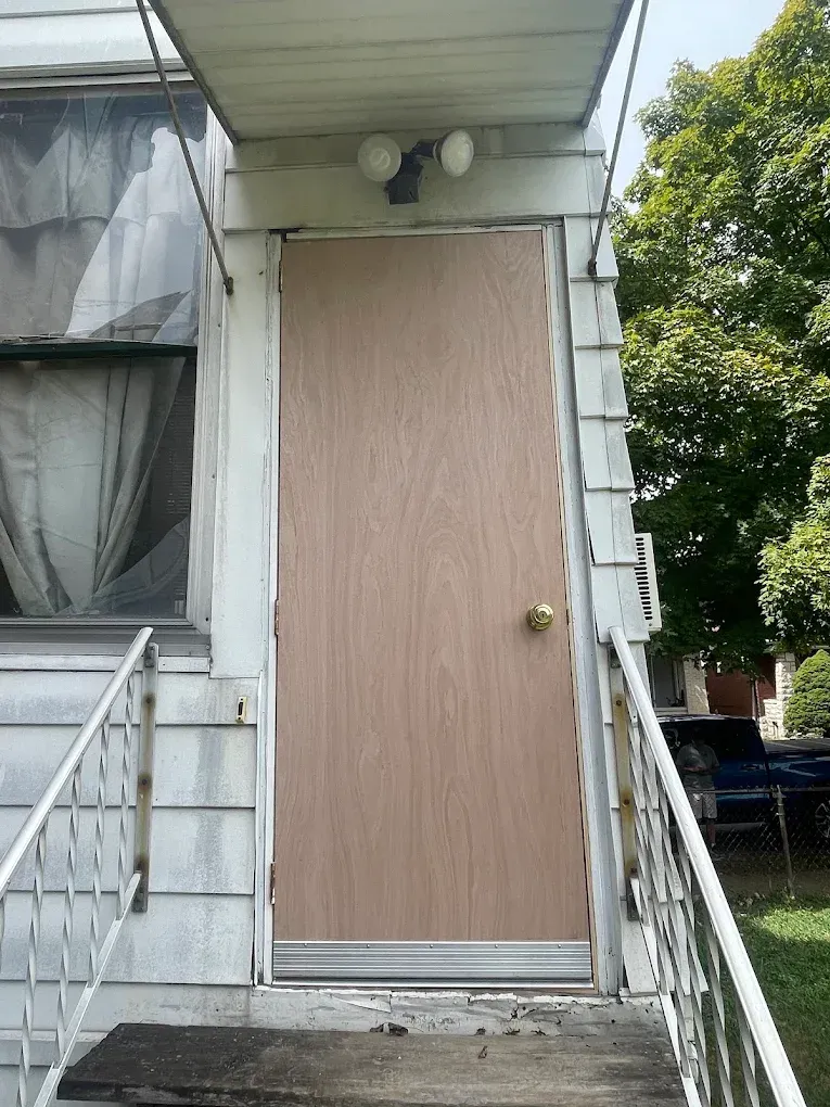 Wooden door on a porch, white siding, metal railing, overhead lights, steps leading up.