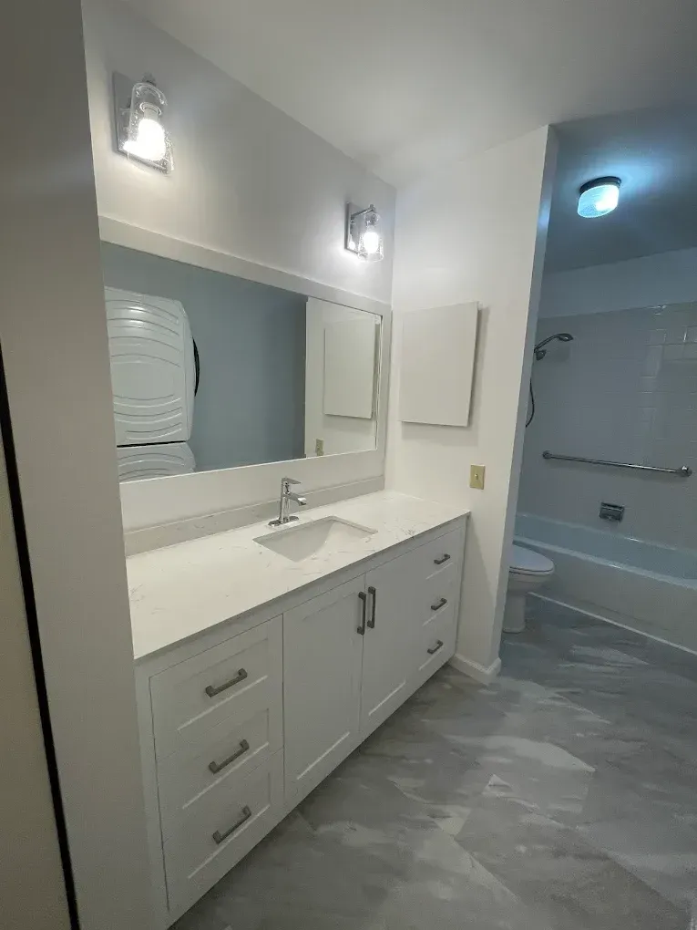White bathroom with a vanity, mirror, and access to a bathtub. Silver fixtures, gray floor, and a white appliance visible.