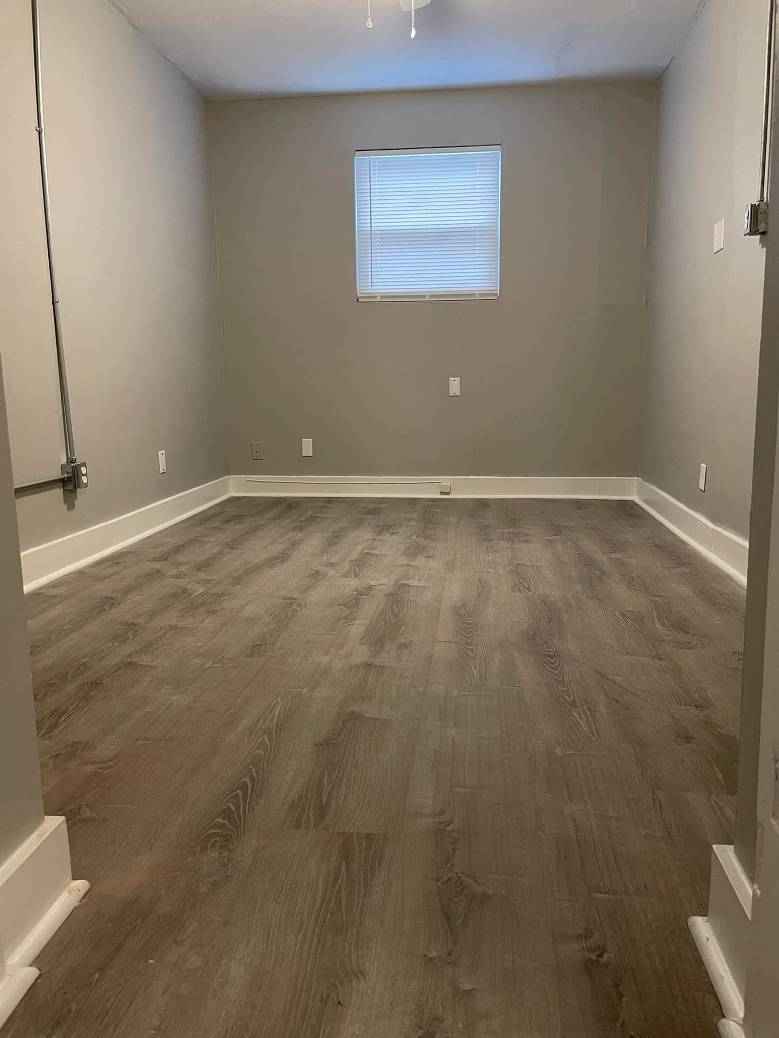 Empty room with gray walls, wood-look floor, small window. Electrical conduit along the left wall.