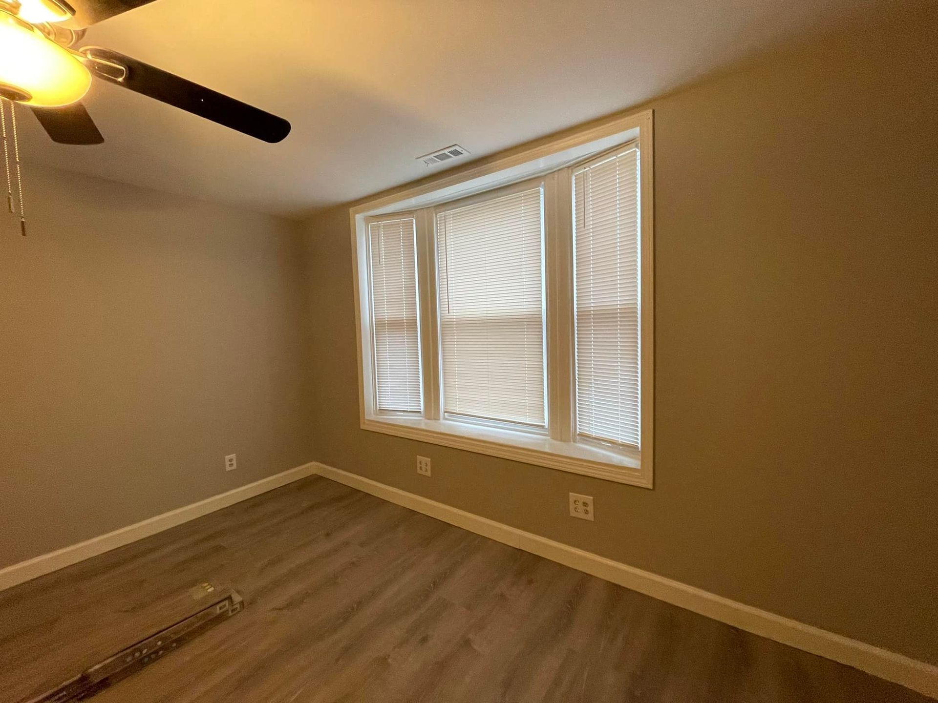 Empty room with window and blinds. Tan walls, wood-look floor, and ceiling fan.