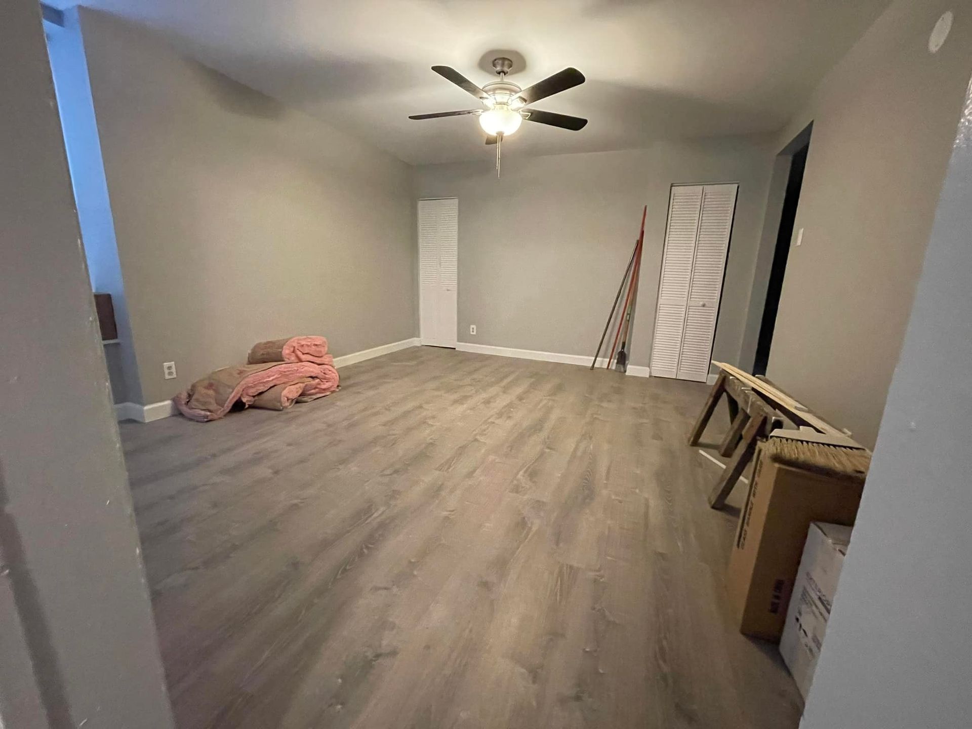 Empty room with gray walls, wood-look flooring, ceiling fan, and construction materials.