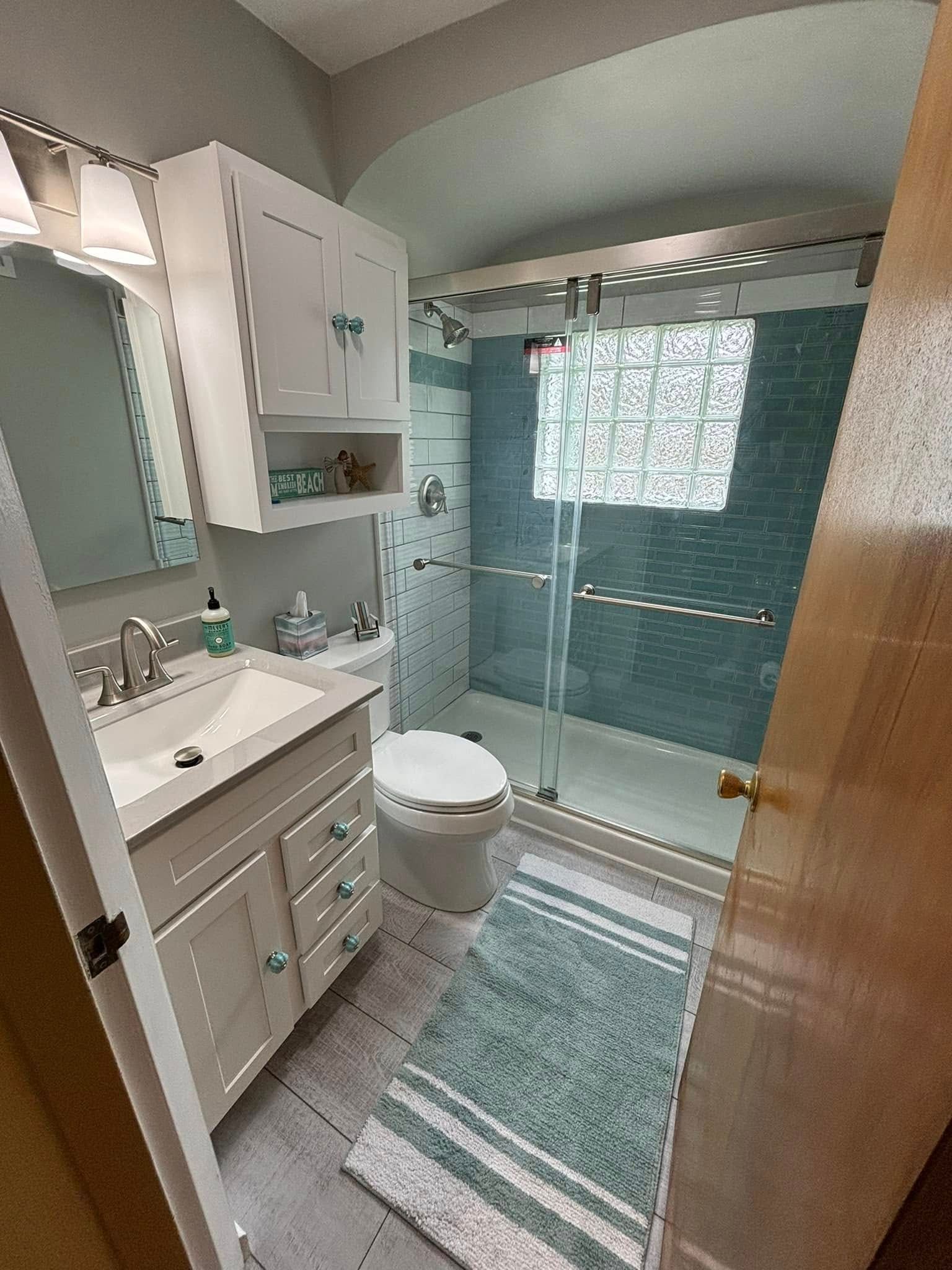 Bathroom with white vanity, toilet, and teal tiled shower. Striped rug on floor.