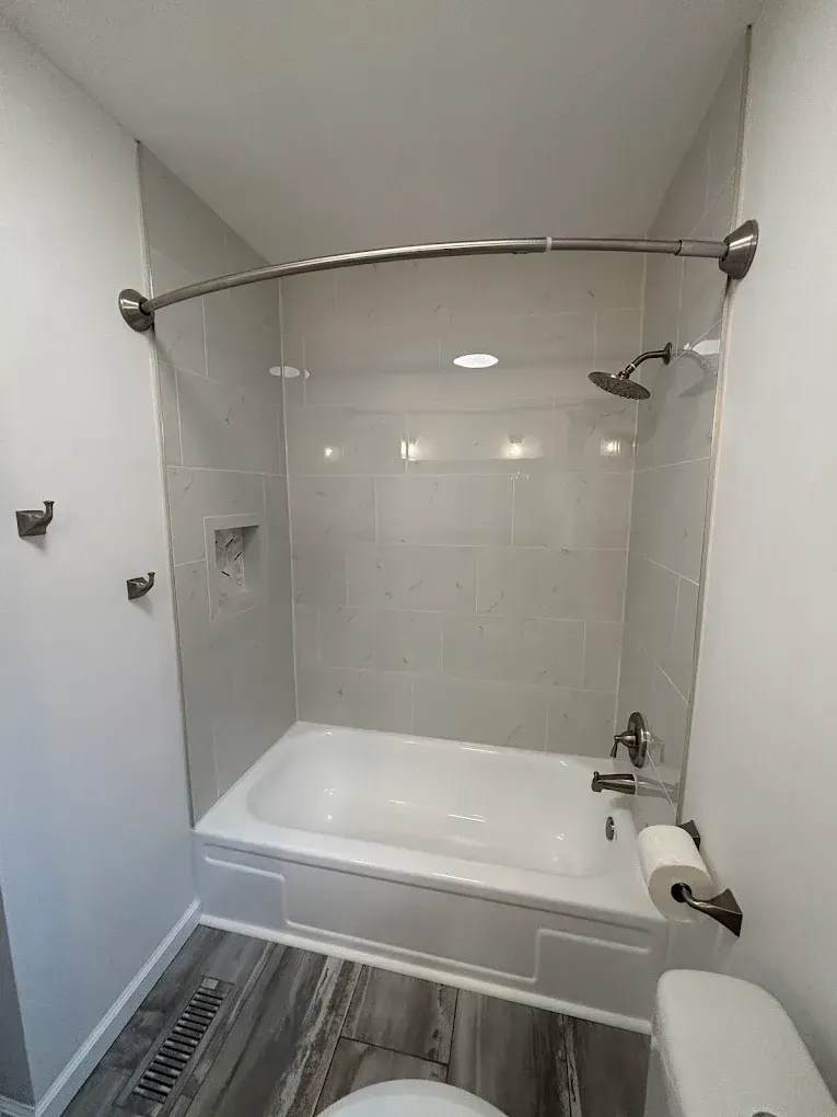 Small bathroom with white tub and gray tiled shower surround, gray and wood flooring, toilet on the right.