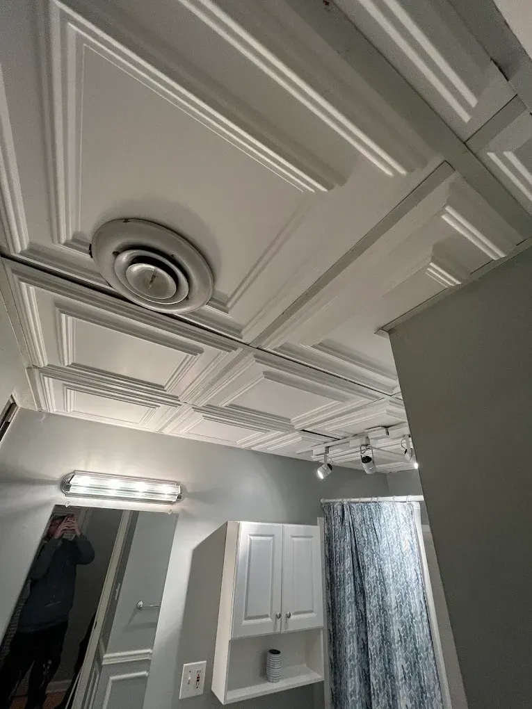 In a bathroom with a white decorative ceiling, white cabinets, and a gray wall, a person is reflected in a mirror.