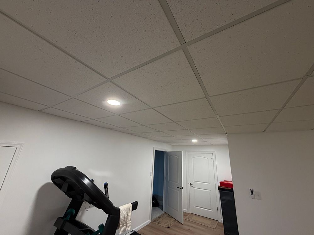 A basement room with white walls, a drop ceiling with recessed lighting, and a treadmill in the foreground.