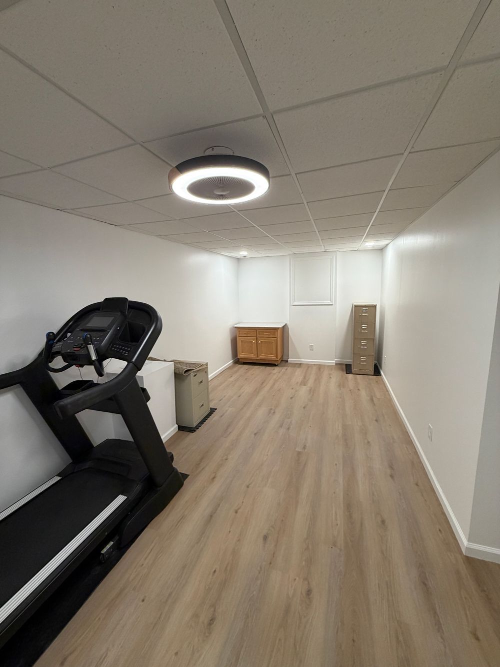 A basement gym with a treadmill, light wood-look flooring, white walls, a circular ceiling light, and storage cabinets.