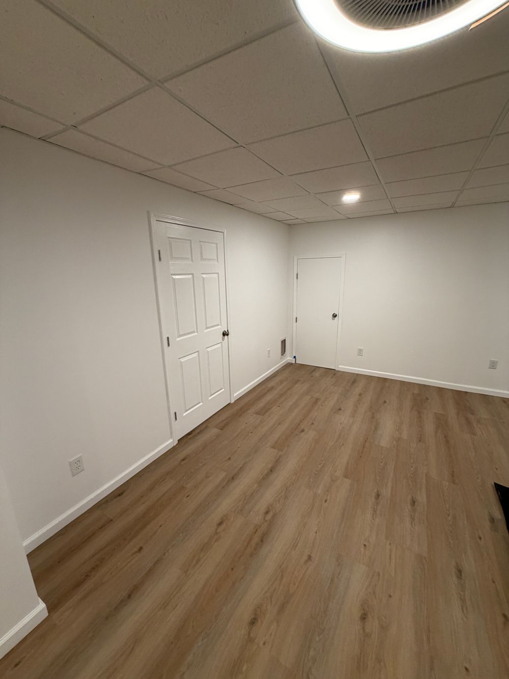 An empty basement room with white walls, a drop ceiling, light wood flooring, and two closed white doors.