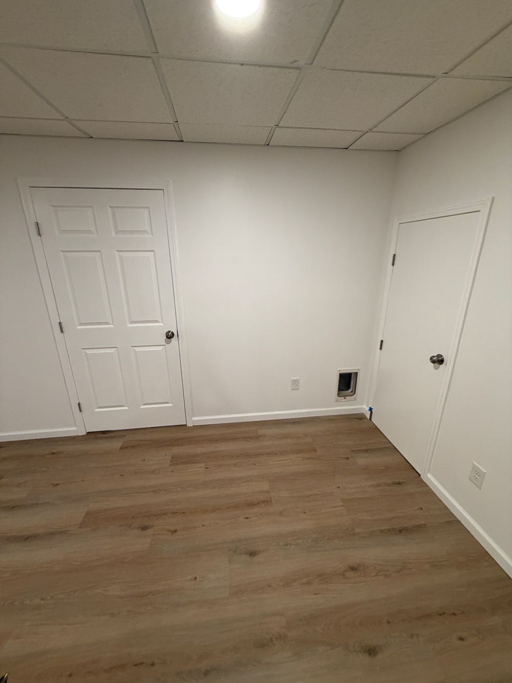 An empty room with white walls, a wood-look floor, a drop ceiling, and two white interior doors.