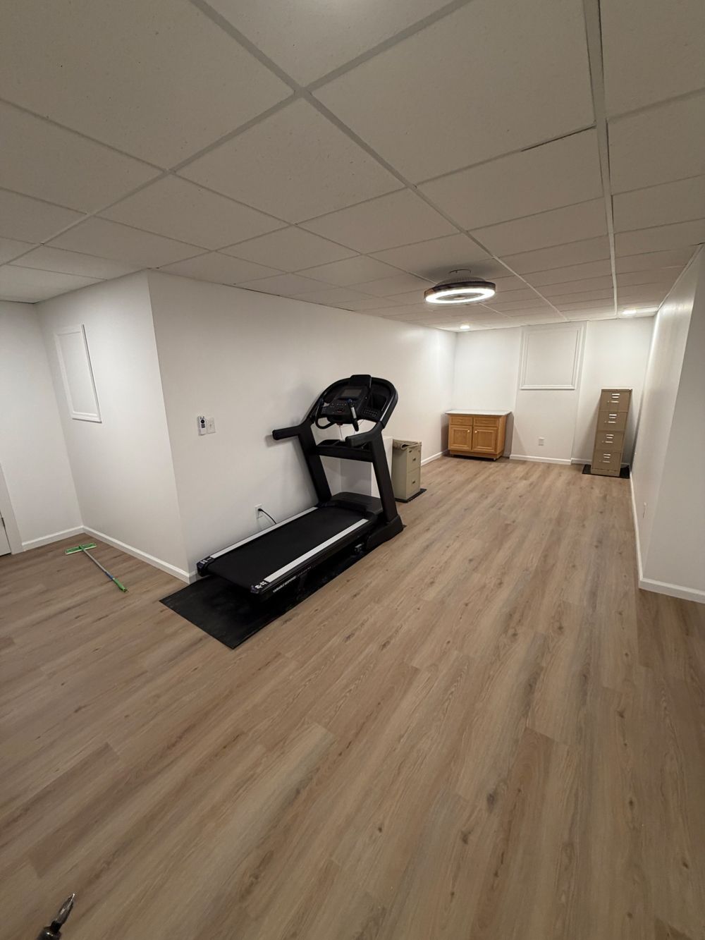 A spacious basement room with light wood flooring, white walls, a treadmill, and a small wooden cabinet.