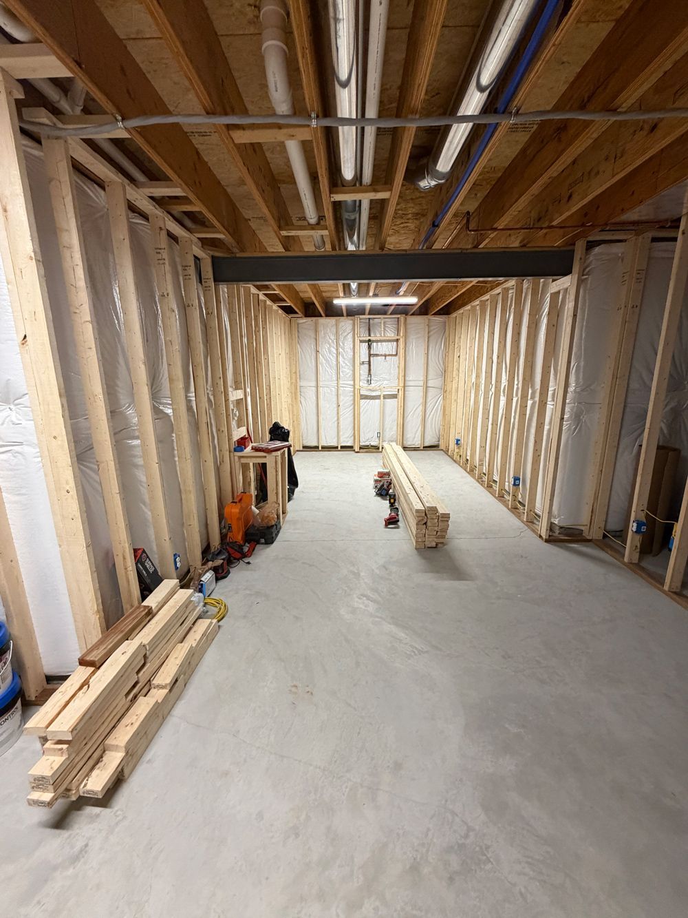 An unfinished basement room with wood-framed walls, exposed ceiling joists, utility pipes, and stacked lumber on the floor.