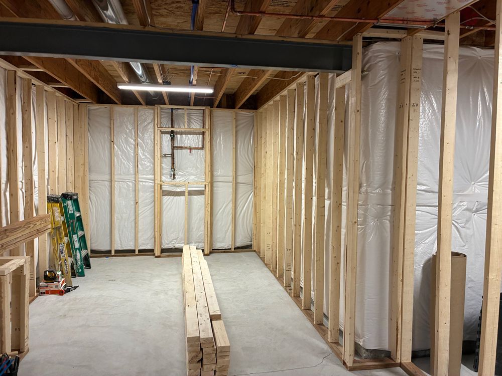 An unfinished basement featuring wooden stud framing, a concrete floor, and plastic-wrapped walls under a steel beam.