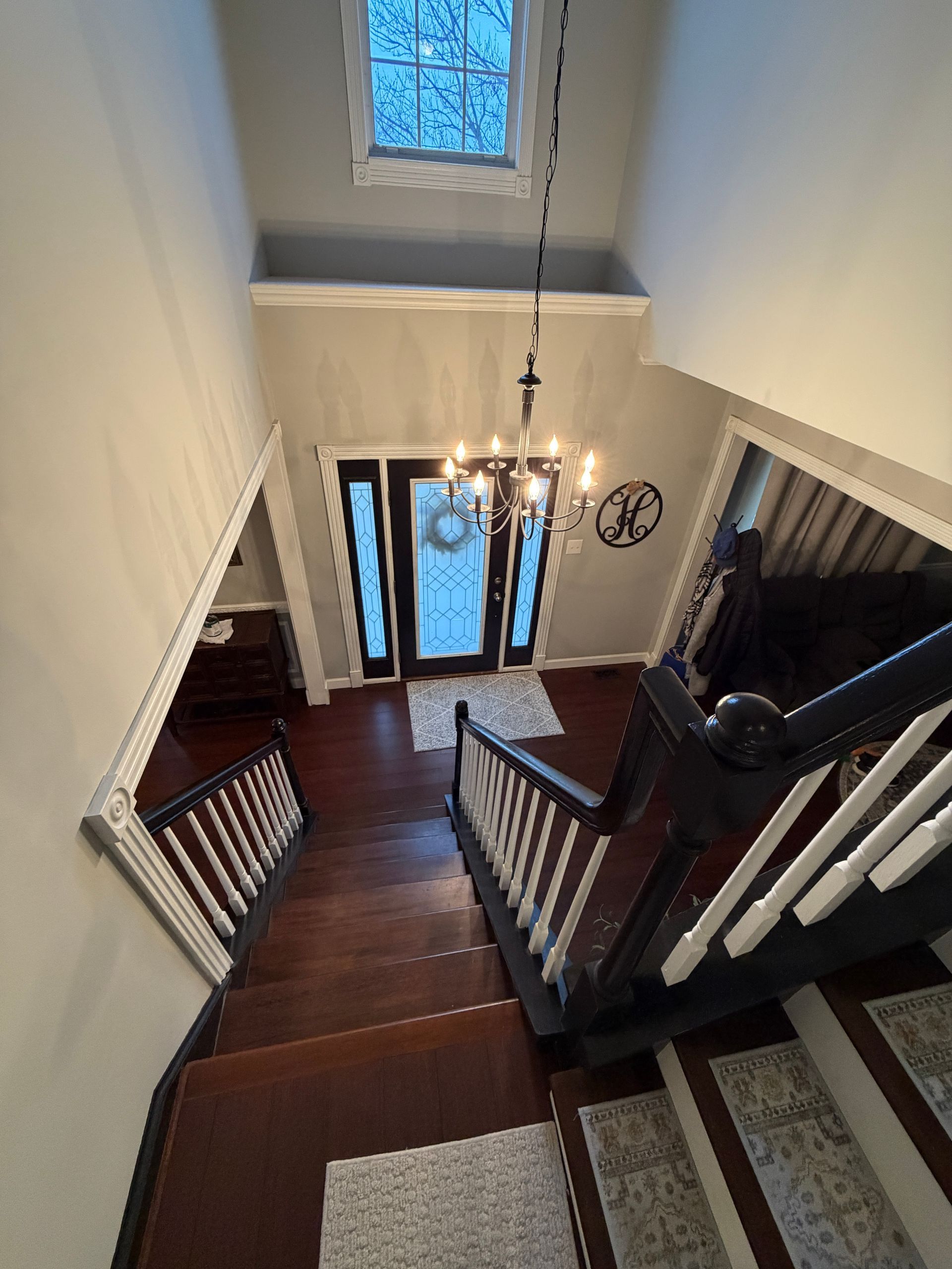 A high-angle view looking down a wooden staircase in an entryway with white walls, a crystal chandelier, and front door.