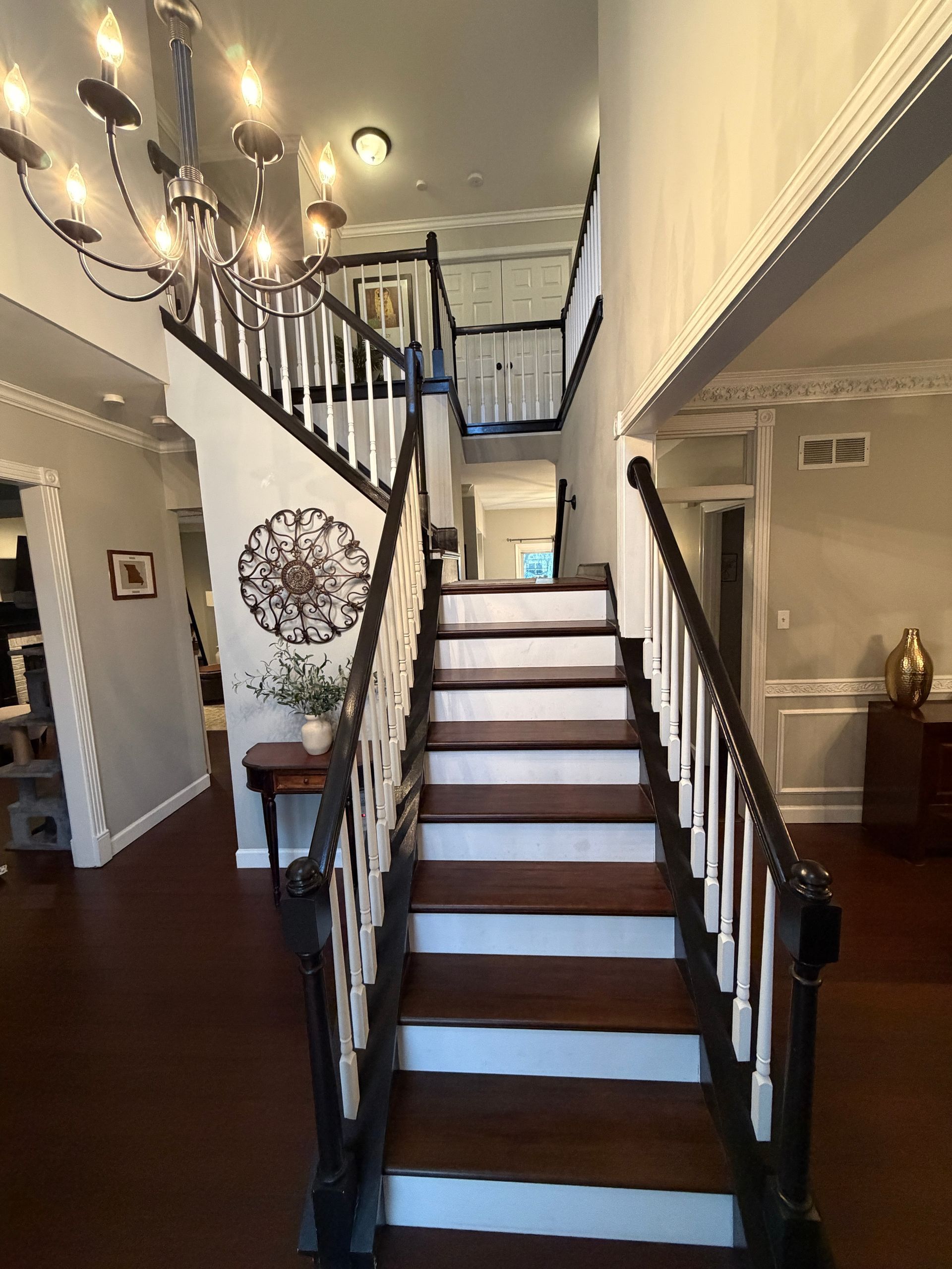 A staircase with dark wooden treads and white risers leads to a landing under a large, modern chandelier.