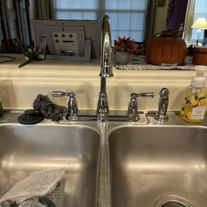 Stainless steel kitchen sink with faucet, handles, and soap dispenser. Pumpkin and window in the background.