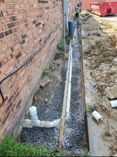 Construction site: trench next to brick wall, containing pipes and gravel. Person in background.
