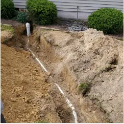 A trench dug in a yard revealing white pipes; shrubs in background.