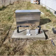 Stainless steel box mounted on concrete, pipes, valves, and wiring outside near a building.