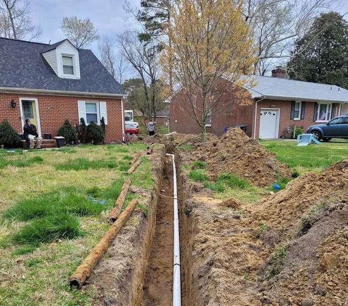 Trench dug in a yard with white pipe installed; piles of dirt, houses in background.