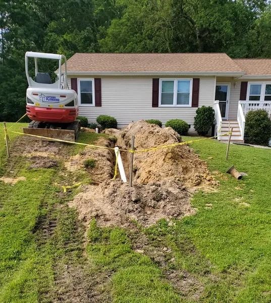 An excavator digs a trench in front of a tan house. Dirt pile and yellow caution tape present.
