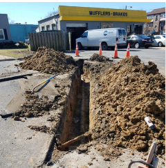 Road construction: Open trench, piles of dirt, orange cones, vehicle repair shop in background.